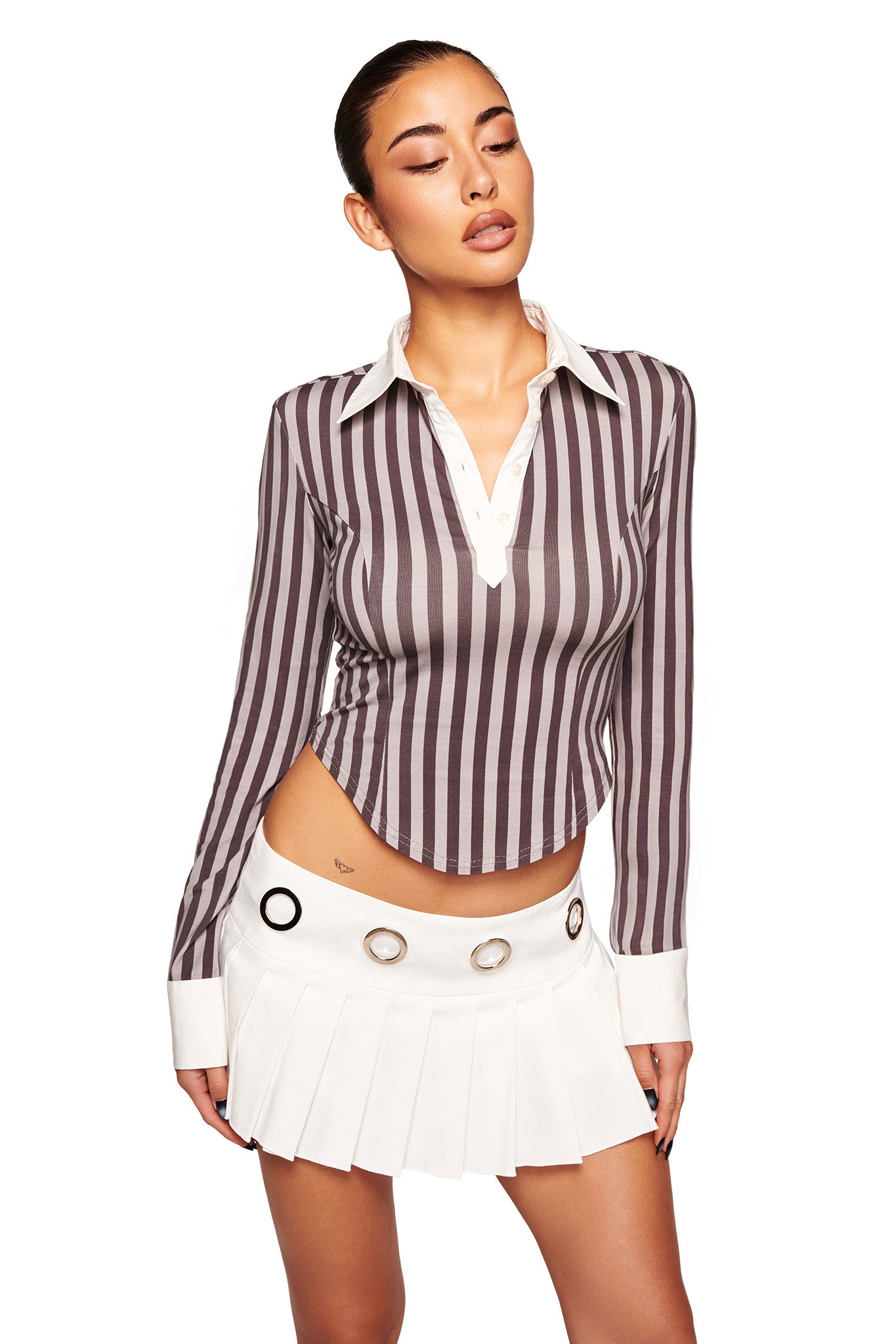 A woman stands against a white background wearing the I.AM.GIA JAYMES SKORT in white and a long-sleeved, brown and white vertically striped shirt with a white collar. The skort features metallic grommet details and a low-rise waist.