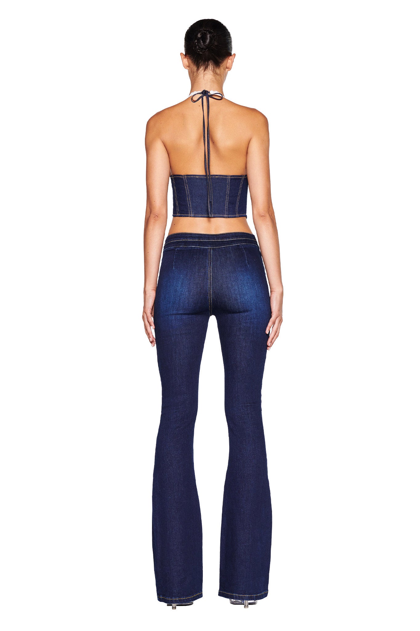 A model is photographed from behind wearing the I.AM.GIA ELLENA CORSET - BLUE : DARK WASH and matching high-waisted flared jeans. The corset features a tie closure at the neck, and the stretch denim jeans have visible stitching and a fitted style through the thighs. The model's hair is in a bun.