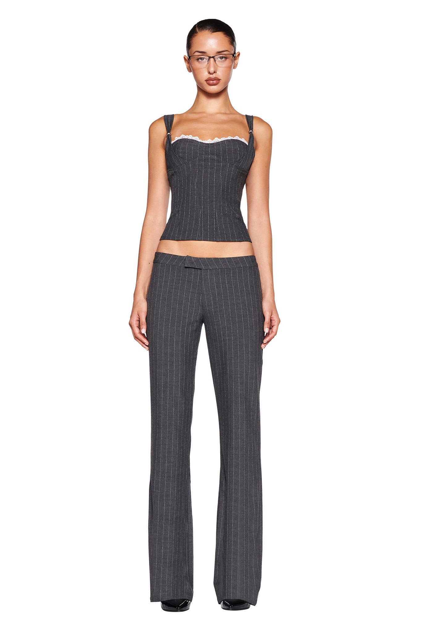A person stands facing forward wearing the MARRION CORSET - GREY : PINSTRIPE by I.AM.GIA, featuring shoulder straps and paired with matching high-waisted, flared pants. They have glasses and dark hair pulled back. The tailored silhouette is accentuated against the plain white background.