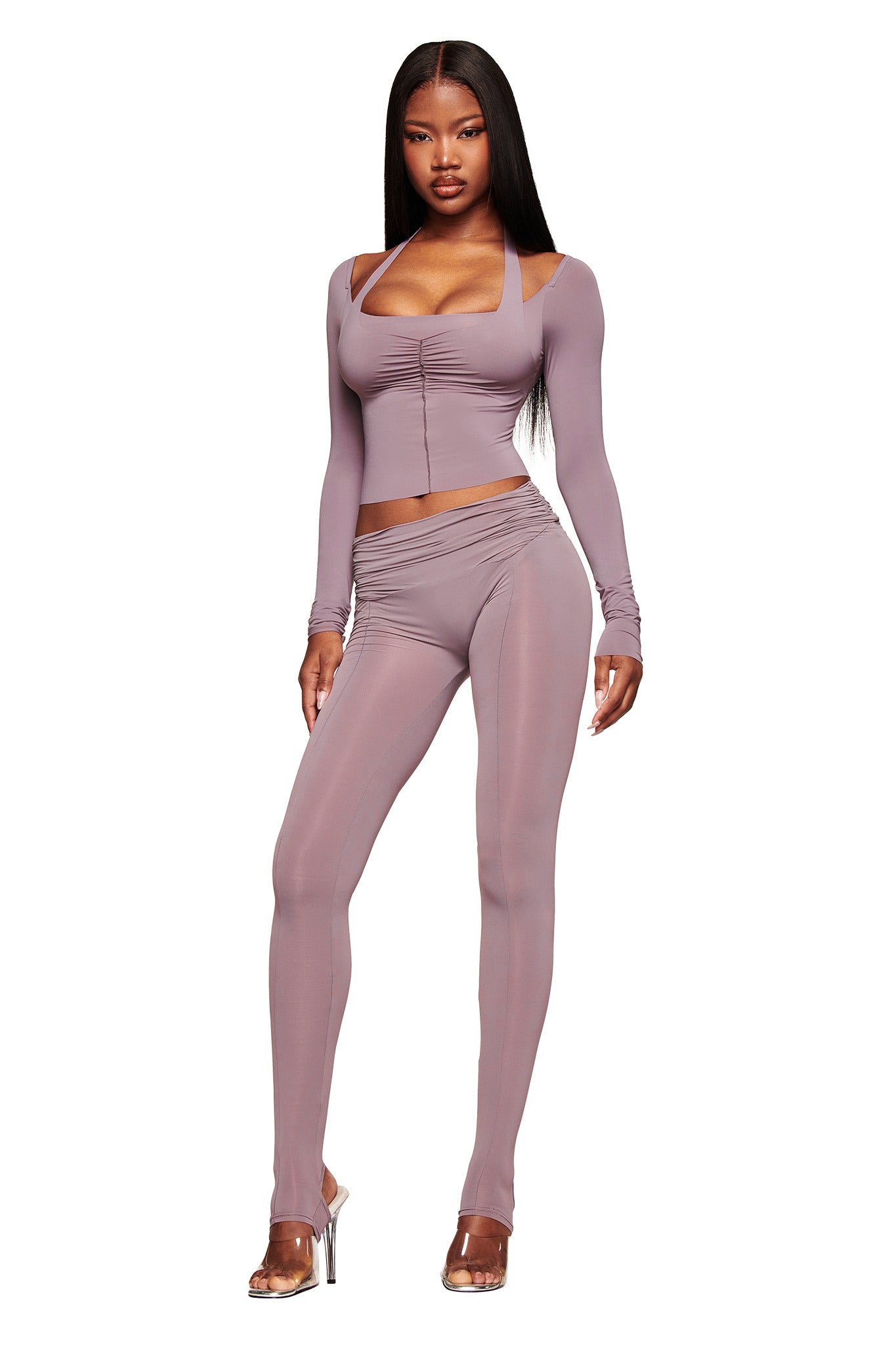 A woman poses confidently in the I.AM.GIA NERINA LONG SLEEVE TOP in dusty lilac, featuring a bodycon fit and square neckline. She pairs it with matching high-waisted leggings and clear heels, her long straight hair flowing against a white backdrop.