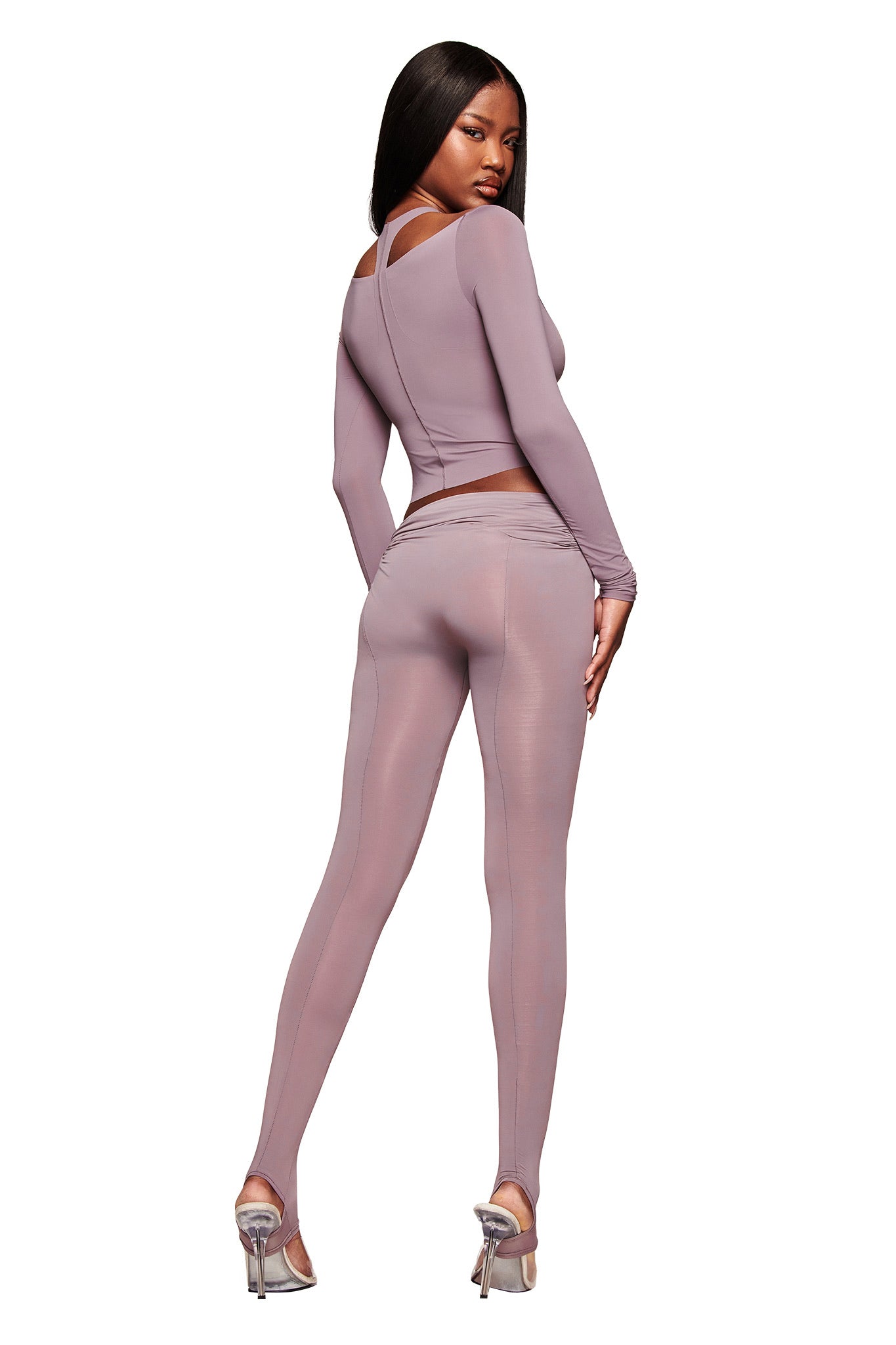 A woman with long straight hair poses in the I.AM.GIA NERINA LEGGING in Dusty Lilac, featuring a fitted, full-length style. She faces away, looking over her shoulder in silver heels against a white background.