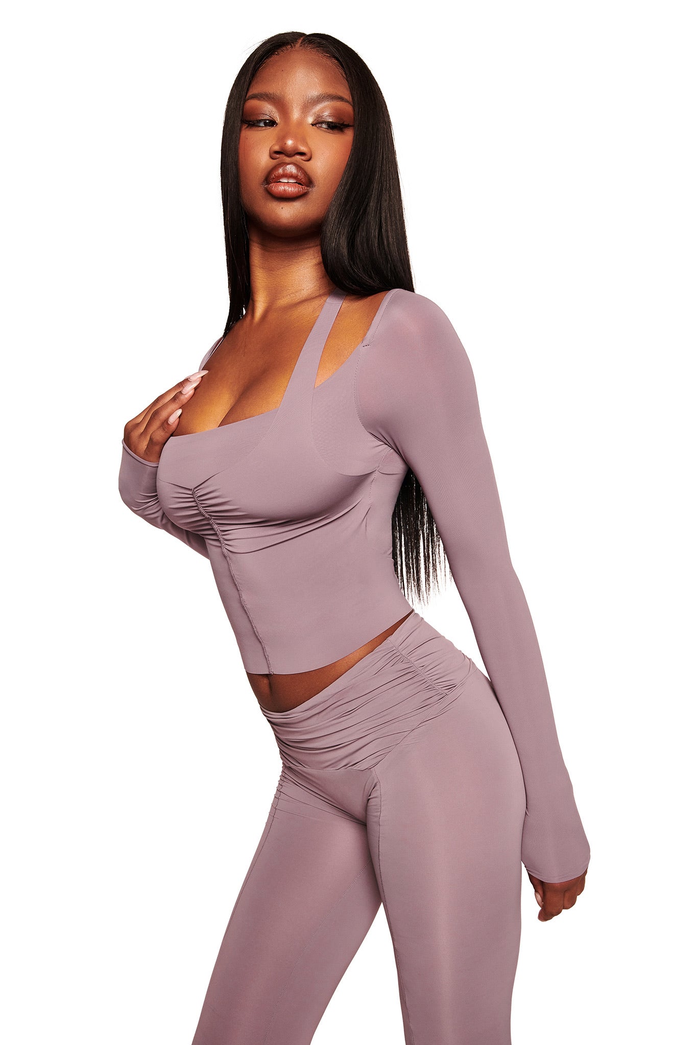 A woman with long straight hair poses confidently in I.AM.GIA’s NERINA LEGGING in Dusty Lilac, styled with a matching, fitted long-sleeve top featuring unique straps, both crafted from soft hosiery fabric, against a white background.