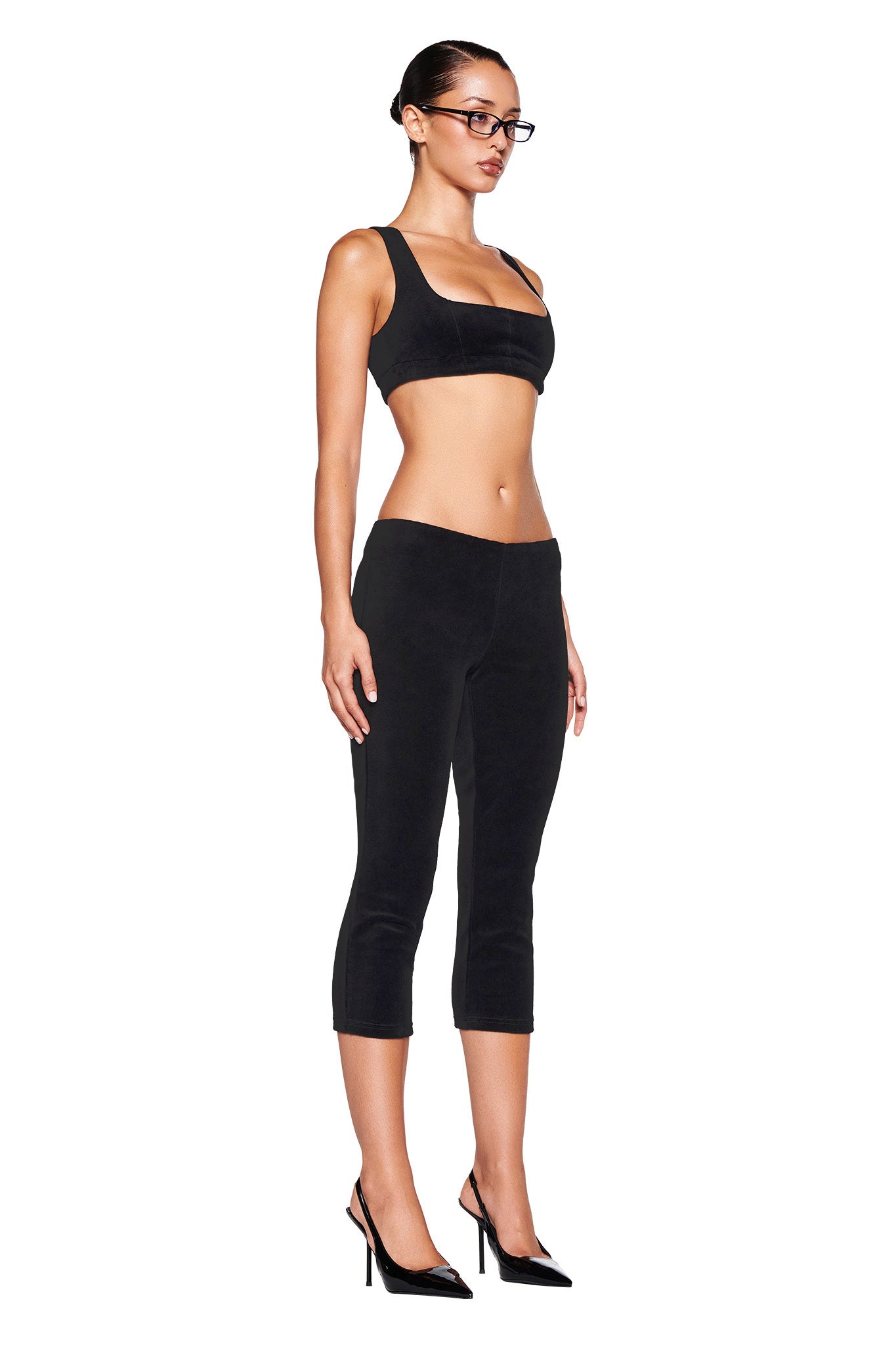 A woman stands against a white background, showcasing the I.AM.GIA LAURA VELVET LEGGING in black. She pairs these form-fitting capri leggings with a matching black crop top and stylish black high-heeled shoes. Her look is completed with a pair of glasses, and her hair is neatly pulled back.
