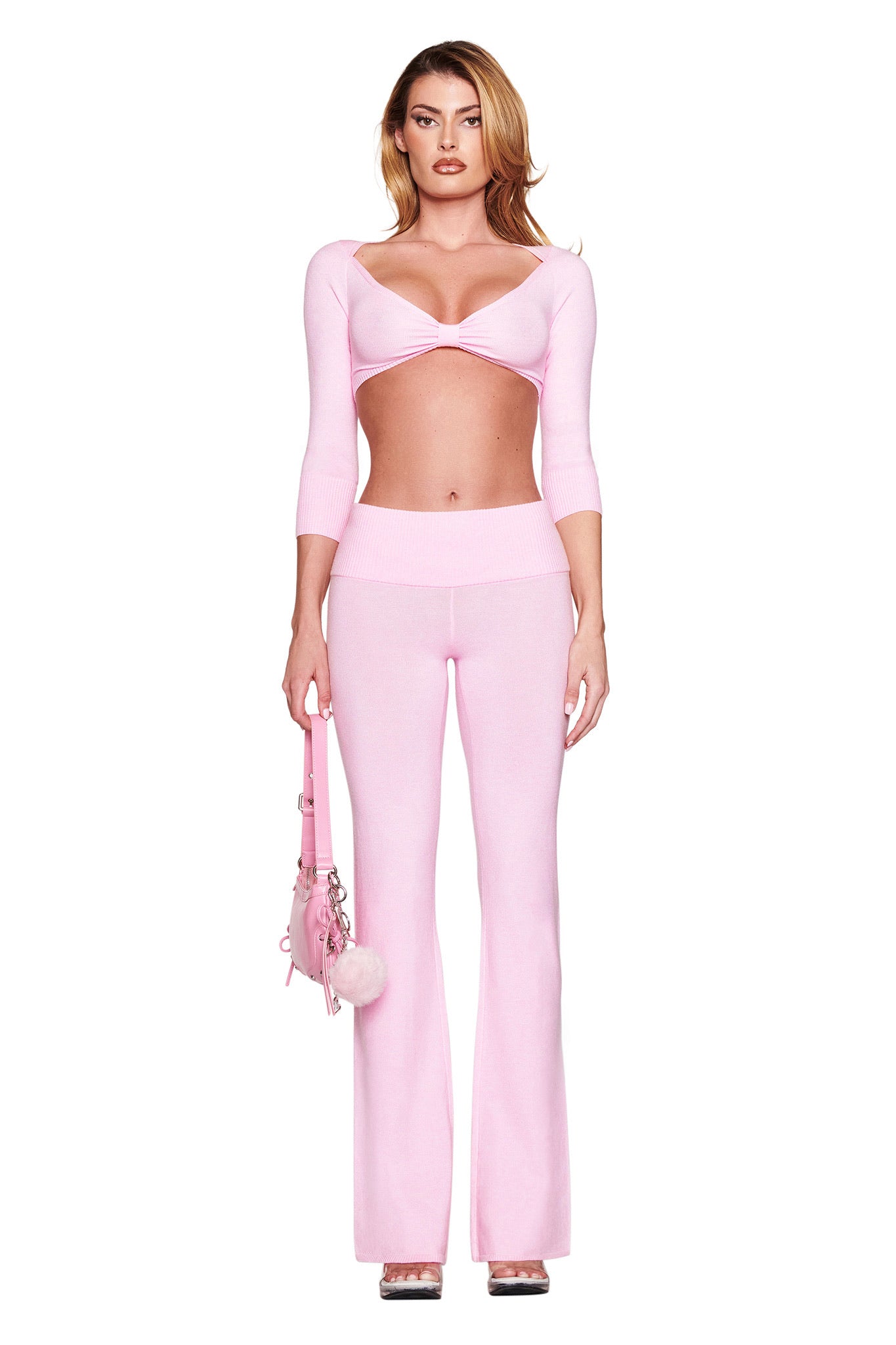 A woman in I.AM.GIA's BABY PINK outfit, featuring a cropped knit top and MORGANA flared high-waisted pants, holds a matching pink handbag with a pom-pom against a white background.