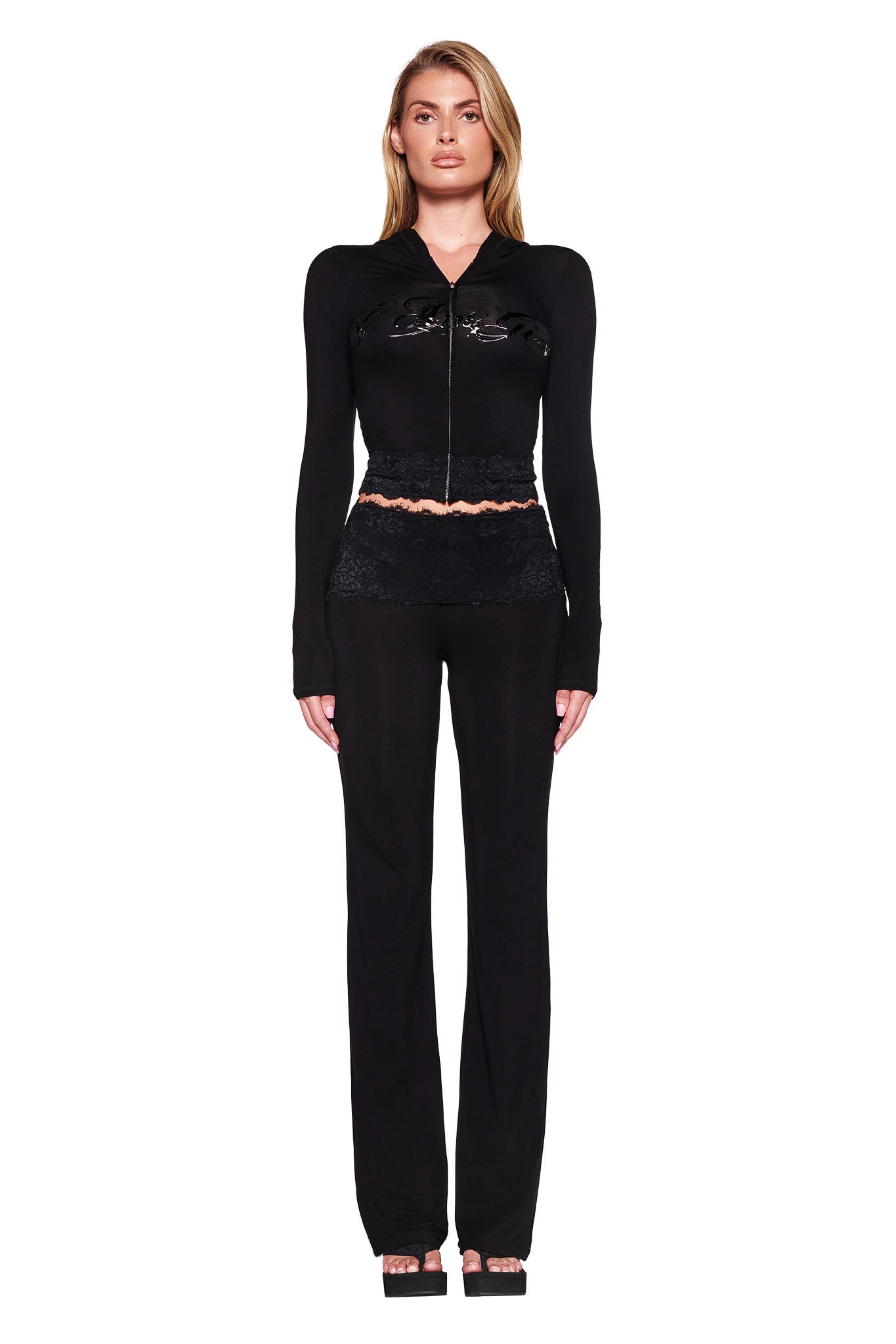 A woman with blonde hair parted to the side is standing against a white background, wearing a black long-sleeve I.AM.GIA TARSHA JACKET - BLACK : TRUE BLACK with a glossy finish on the chest, paired with black pants and black open-toe heels. The jacket features lace detailing at the waistline and prominently displays the I.AM.GIA logo.