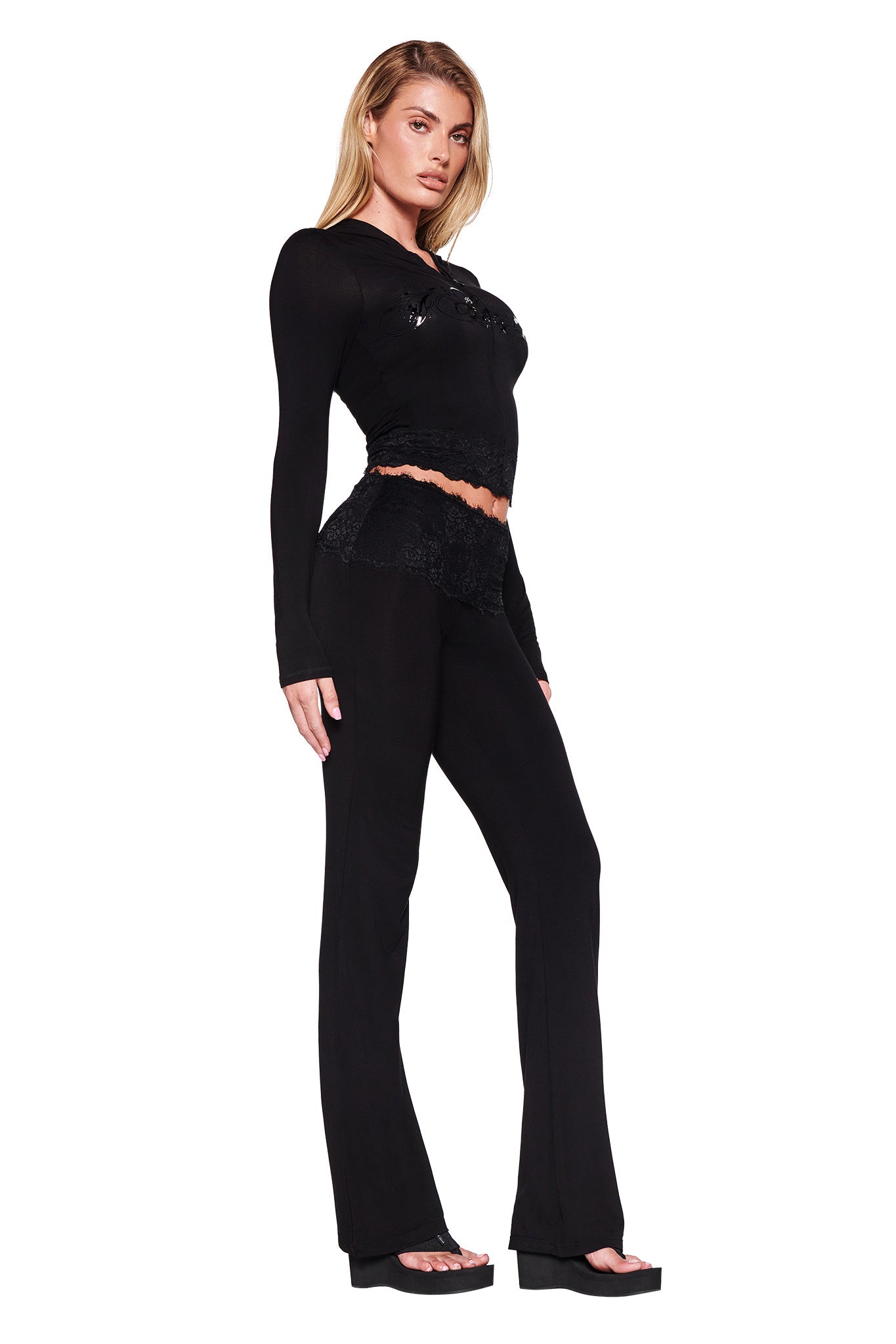 A person with long blonde hair is dressed in a black ensemble, featuring a long-sleeved crop top adorned with lace details and flared pants. They pair the outfit with black platform sandals. Completing the look is the I.AM.GIA TARSHA JACKET - BLACK : TRUE BLACK, resulting in an edgy yet polished appearance against the white background.