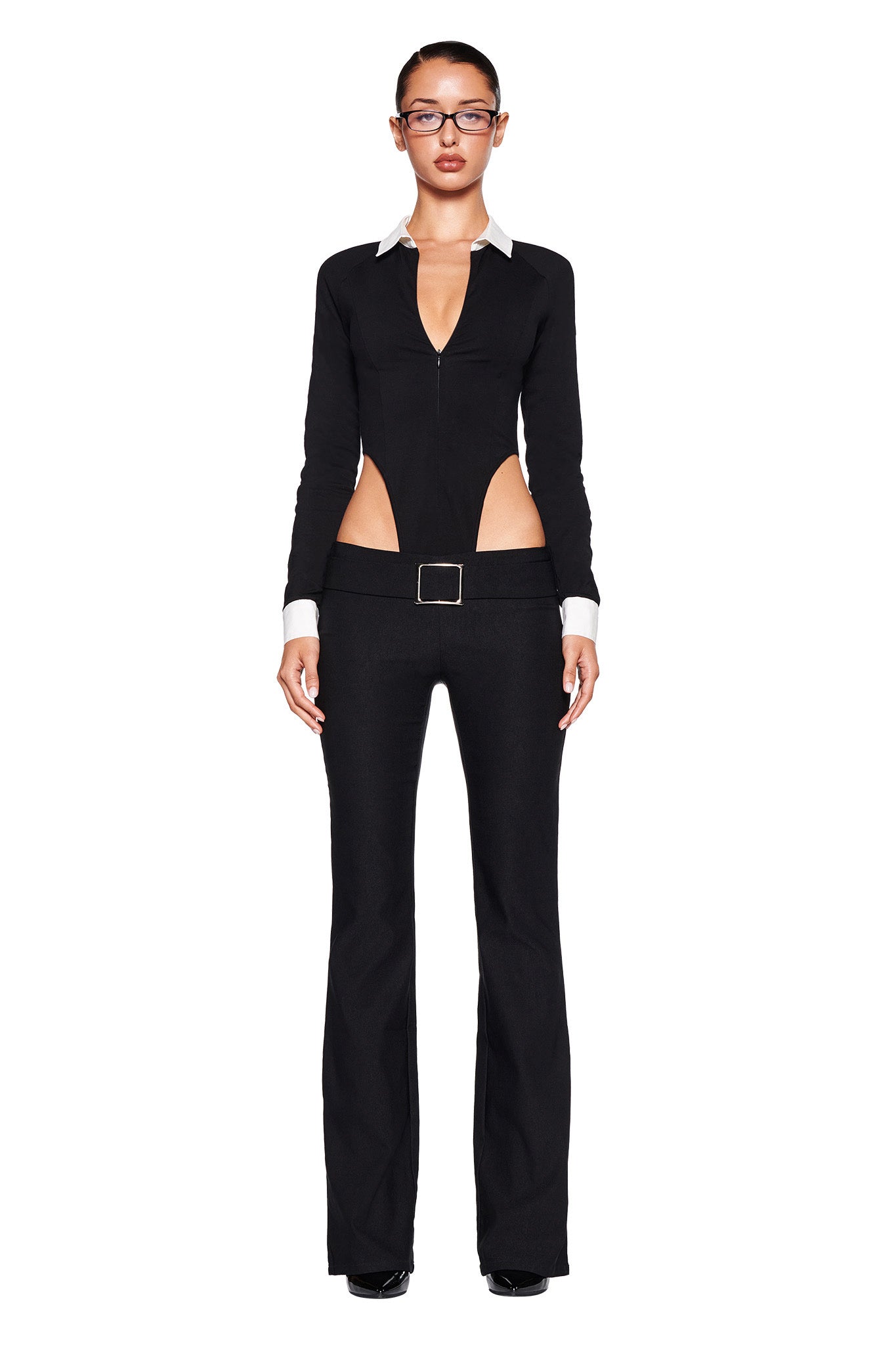 A woman stands against a white background, wearing an I.AM.GIA ensemble featuring a form-fitting top with a plunging neckline, cutout sides, long sleeves with white cuffs, and matching black SHARAH PANT with flared legs. She also wears glasses.