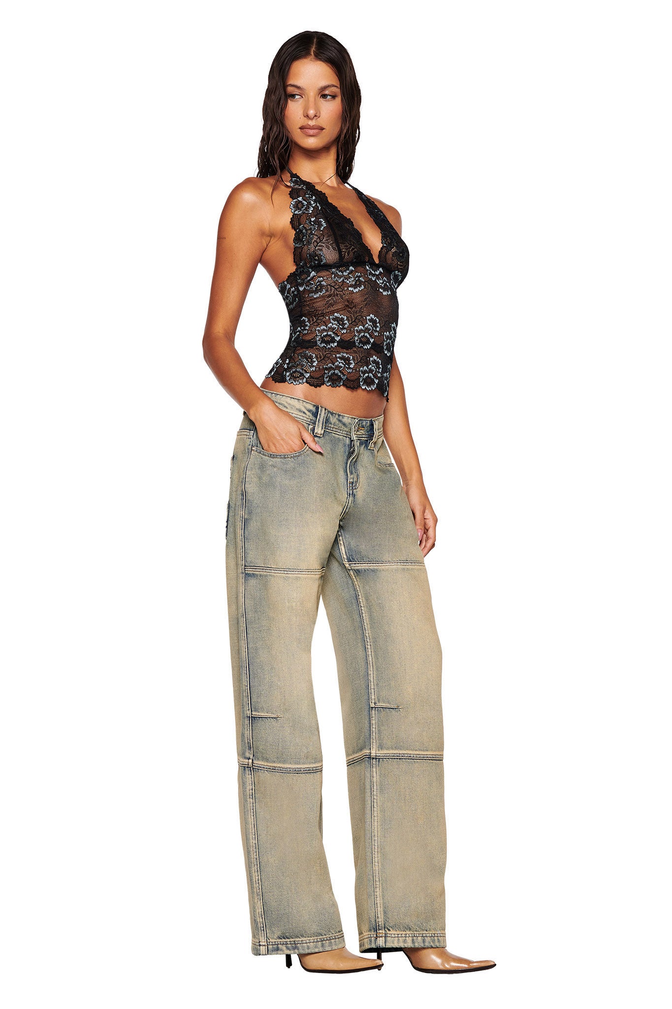 A woman stands confidently, wearing the I.AM.GIA GEM HALTER TOP in black lace fabrication and loose-fitting beige jeans featuring visible stitching. Her hair flows in loose waves, and she completes the look with pointed heel shoes against a plain white background.