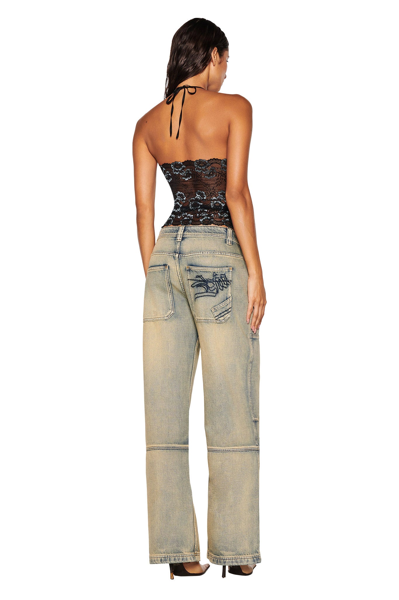 A woman stands with her back to the camera, highlighting the intricate lace design of her I.AM.GIA GEM HALTER TOP in black, paired with wide-legged light blue jeans featuring large pockets and distressed accents. Her long, dark hair flows down as she poses against a plain white backdrop.