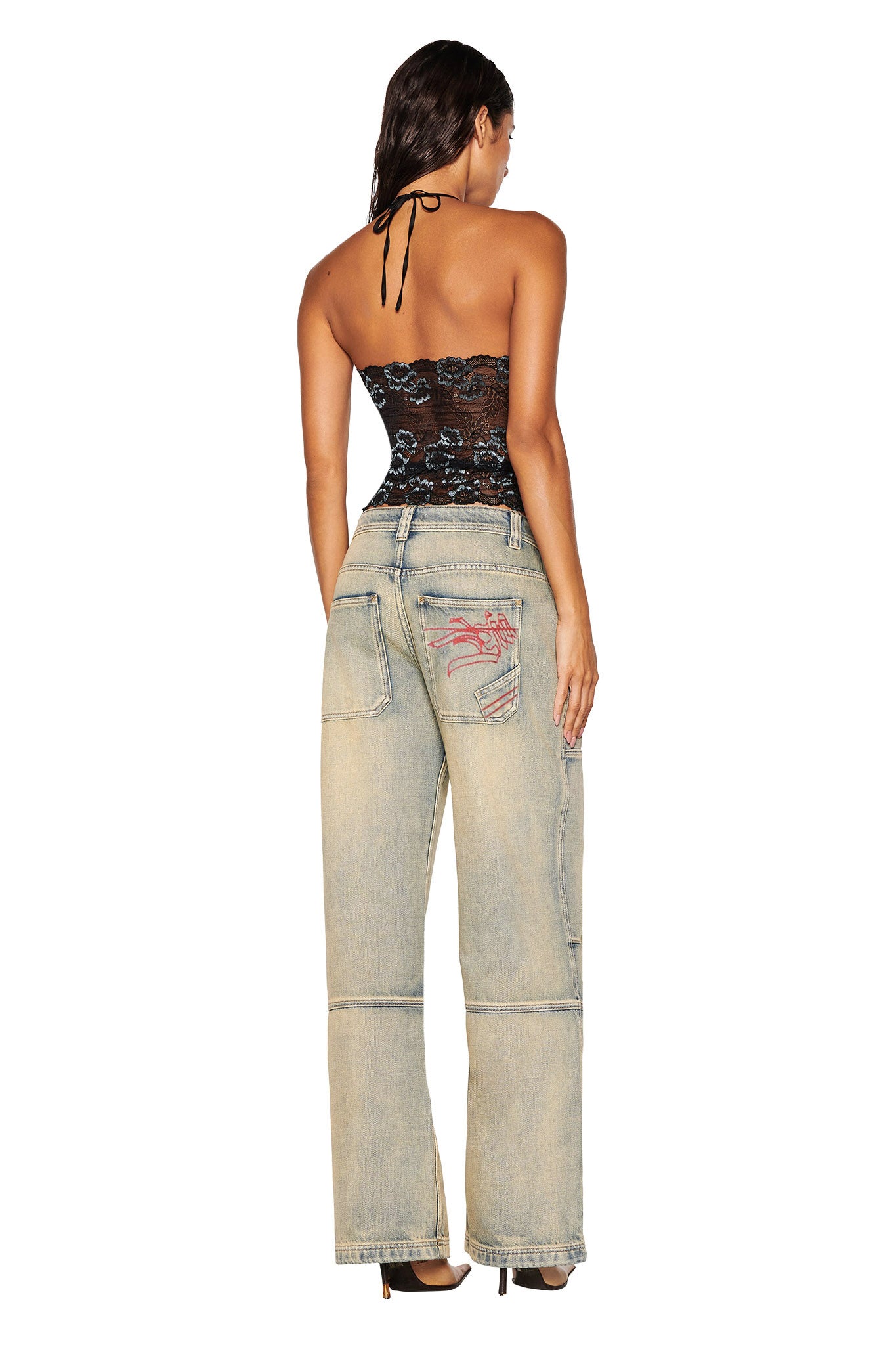 A woman faces away, wearing a black strapless lace top and I.AM.GIA MISTY JEAN - TAN 2.0 with red pocket stitching and a low-rise waist, standing against a plain white background.