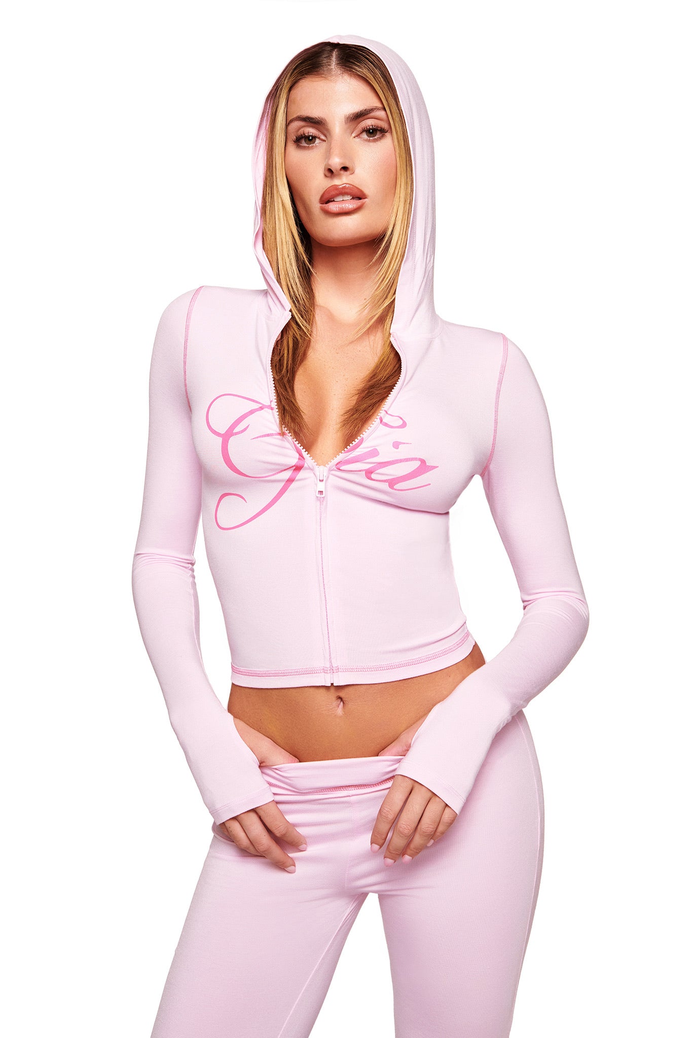 A woman wears a pastel pink I.AM.GIA zip-up hoodie with "Gia" on the chest and matching BLARE TRACKPANT - PINK, featuring a low-rise waist, posing with the hood up against a white background.