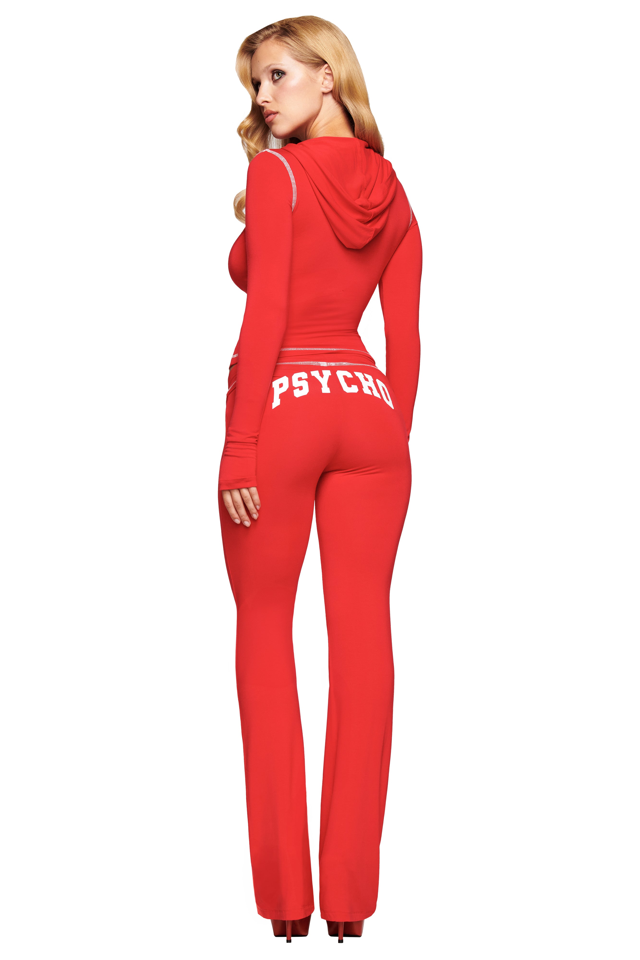A woman with long blonde hair wears I.AM.GIA's red BLARE TRACKPANT - RED : PSYCHO : SALSA, featuring "PSYCHO" in white on the back, as she stands looking over her shoulder.