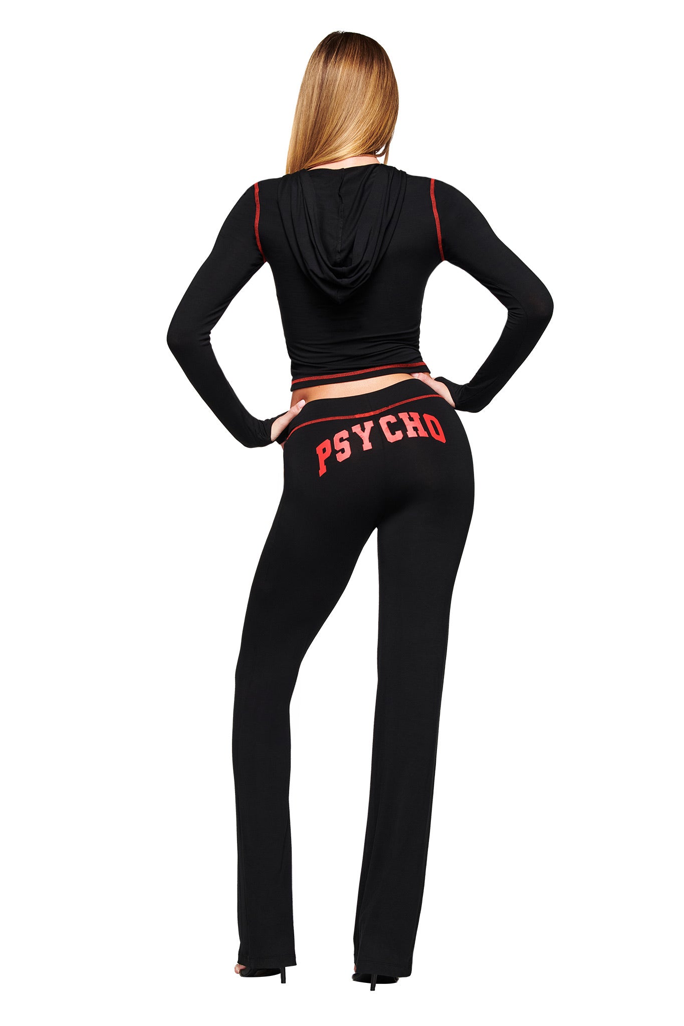 A person with long blonde hair, wearing a black hooded crop top and I.AM.GIA’s BLARE TRACKPANT - BLACK PSYCHO featuring a low-rise waist and bold red "PSYCHO" lettering on the back, stands facing away against a white background.