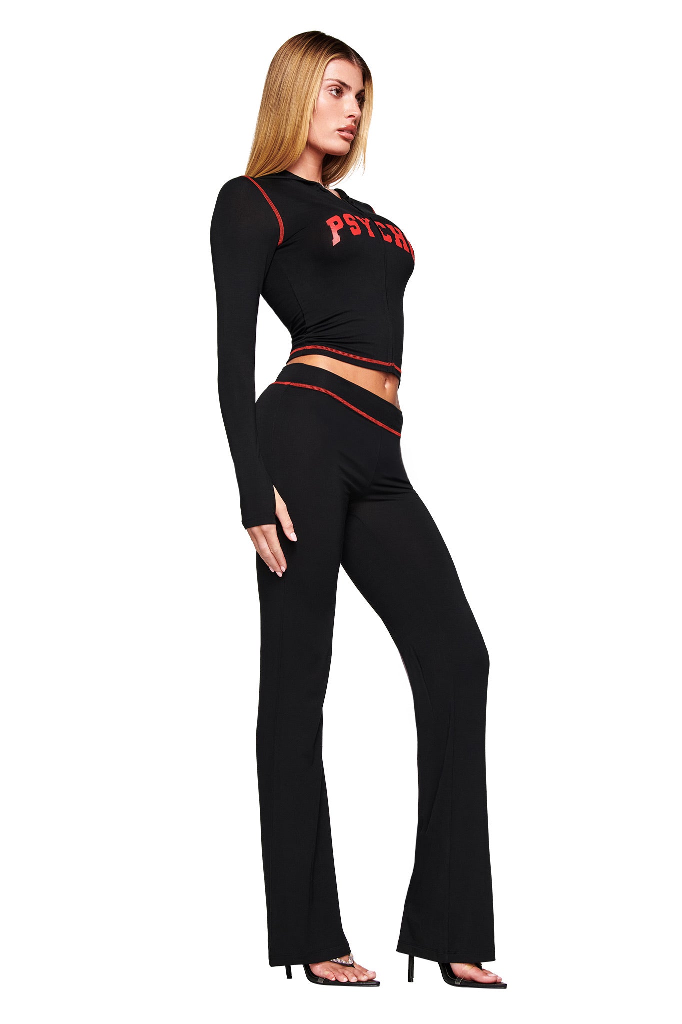 A woman wears the I.AM.GIA BLARE TRACKPANT - BLACK PSYCHO, a black flared pant with red trim and a matching long-sleeve crop top featuring red PSYCHO logo lettering, styled with black high-heeled sandals against a white backdrop.