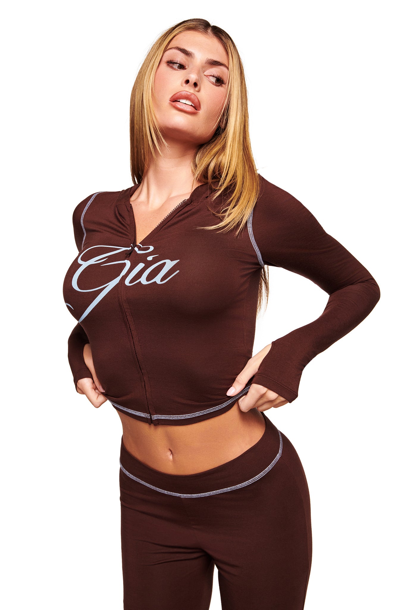 A woman with long blonde hair models the I.AM.GIA BLARE HOODIE in brown with white stitching and "Gia" script, posing with her hands on her waist against a plain white background.