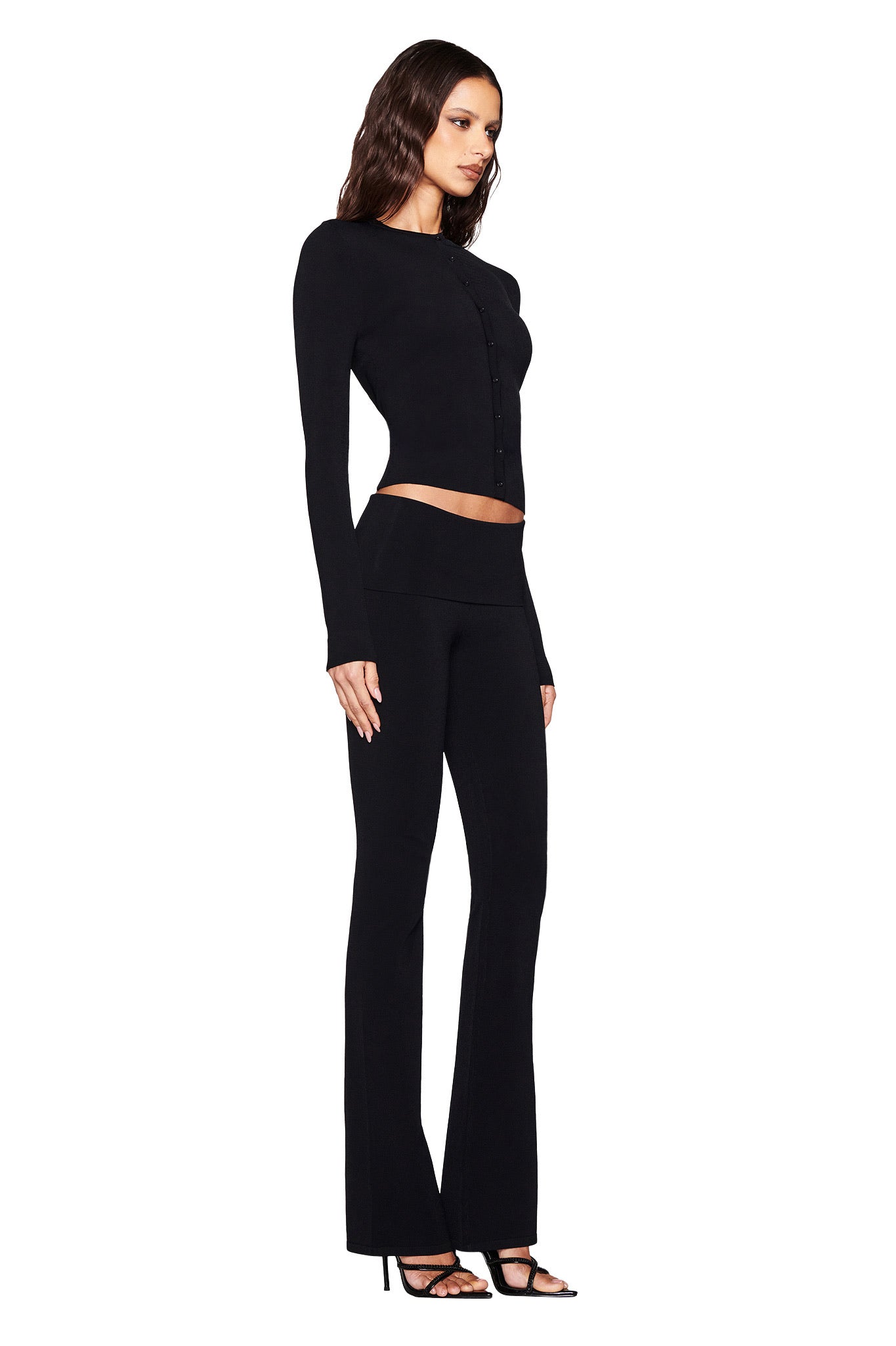 A woman in a fitted black cardigan and AUDRINA PANT 2.0 by I.AM.GIA with a chic black foldover waistband stands in profile against a white background, completing her look with sleek black high-heeled sandals.