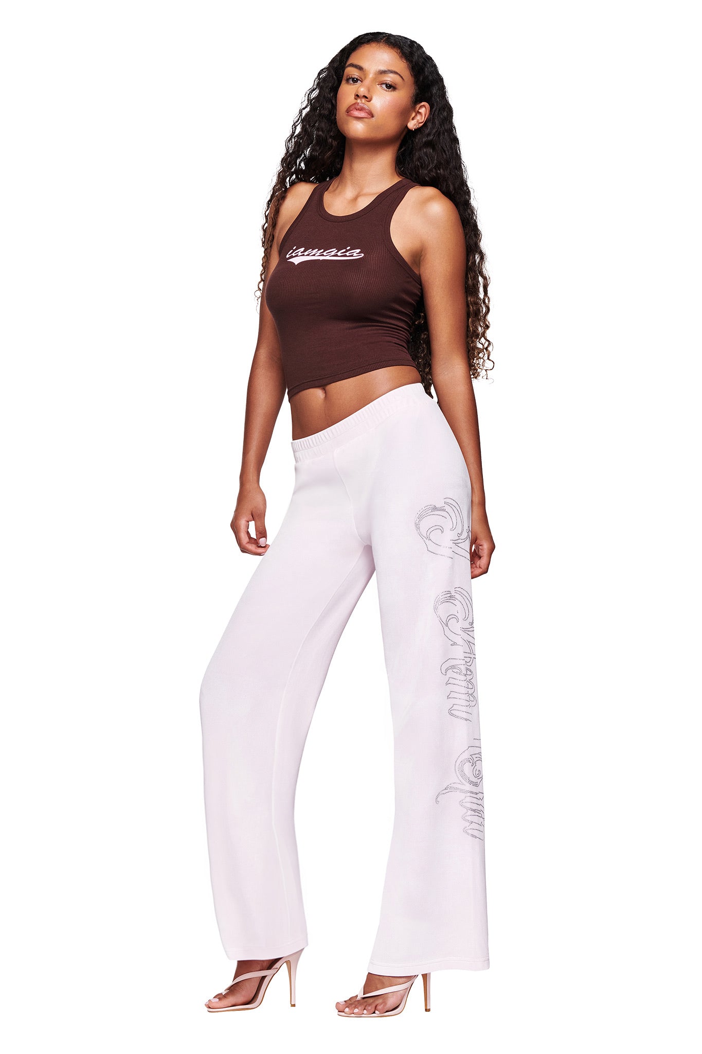 A woman with long curly hair wears the I.AM.GIA PENELOPE TANK in brown, featuring GIA logo artwork, and light pink wide-leg pants with a side design, paired with clear heels, standing against a white background.