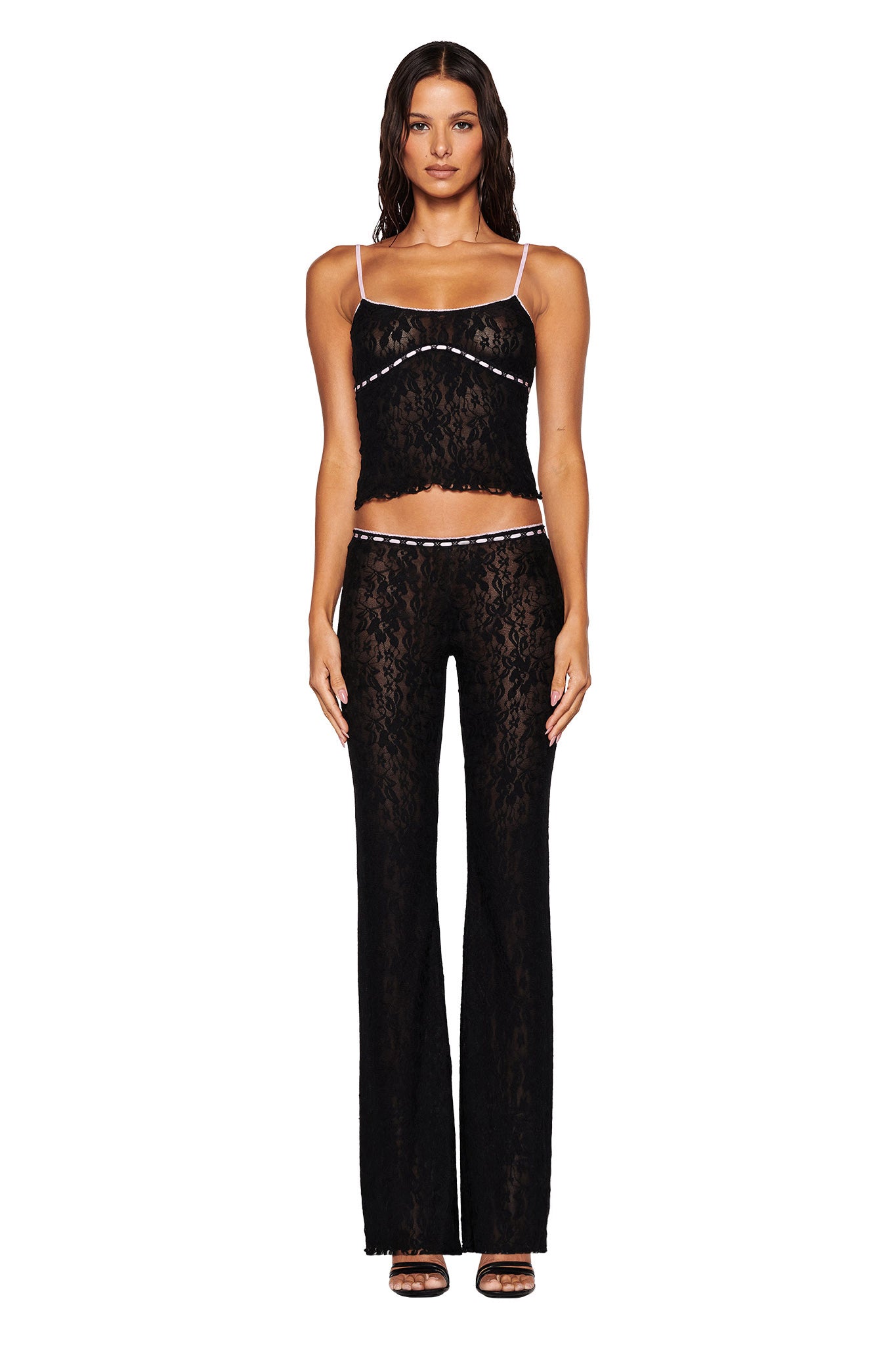A woman stands facing forward in the LORAH TOP - BLACK by I.AM.GIA, featuring a sheer black lace cami with adjustable satin straps, paired with matching pants. The ensemble showcases a bodycon fit that perfectly complements her long, wavy hair and high heels against a plain white background.