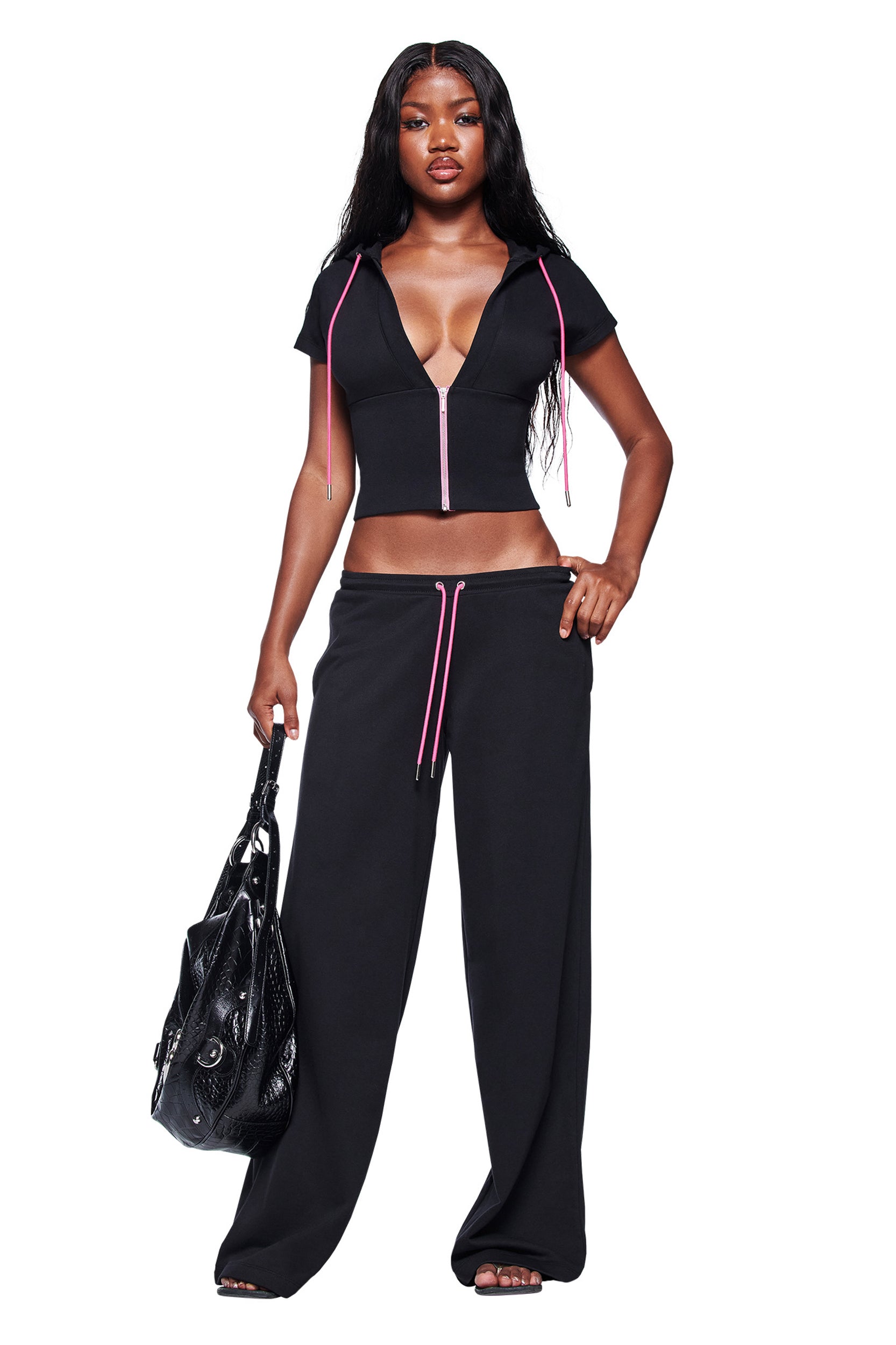 A woman stands confidently against a white background wearing I.AM.GIA’s ARLO TRACK PANT in black with pink drawstrings, paired with a black short-sleeve zip-up V-neck crop top, black sandals, and a large black handbag.
