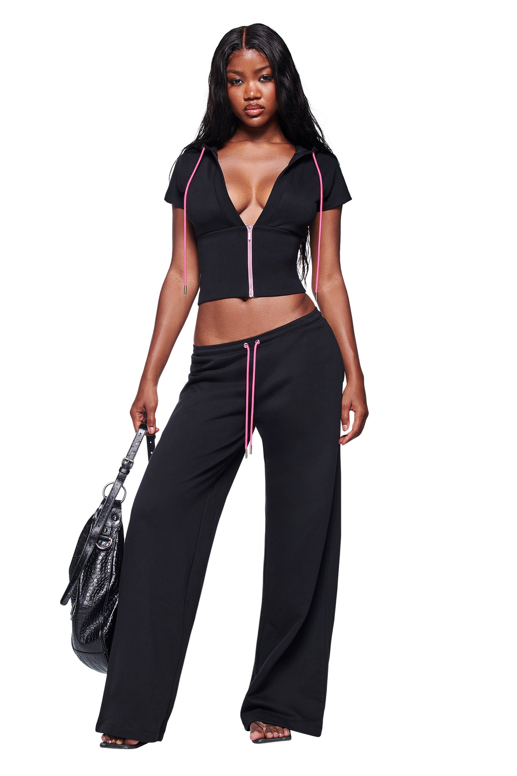 A woman with long black hair wears the I.AM.GIA ARLO TRACK PANT in black, paired with a black, zip-up crop top with pink drawstrings. She confidently holds a large black handbag against a white background.