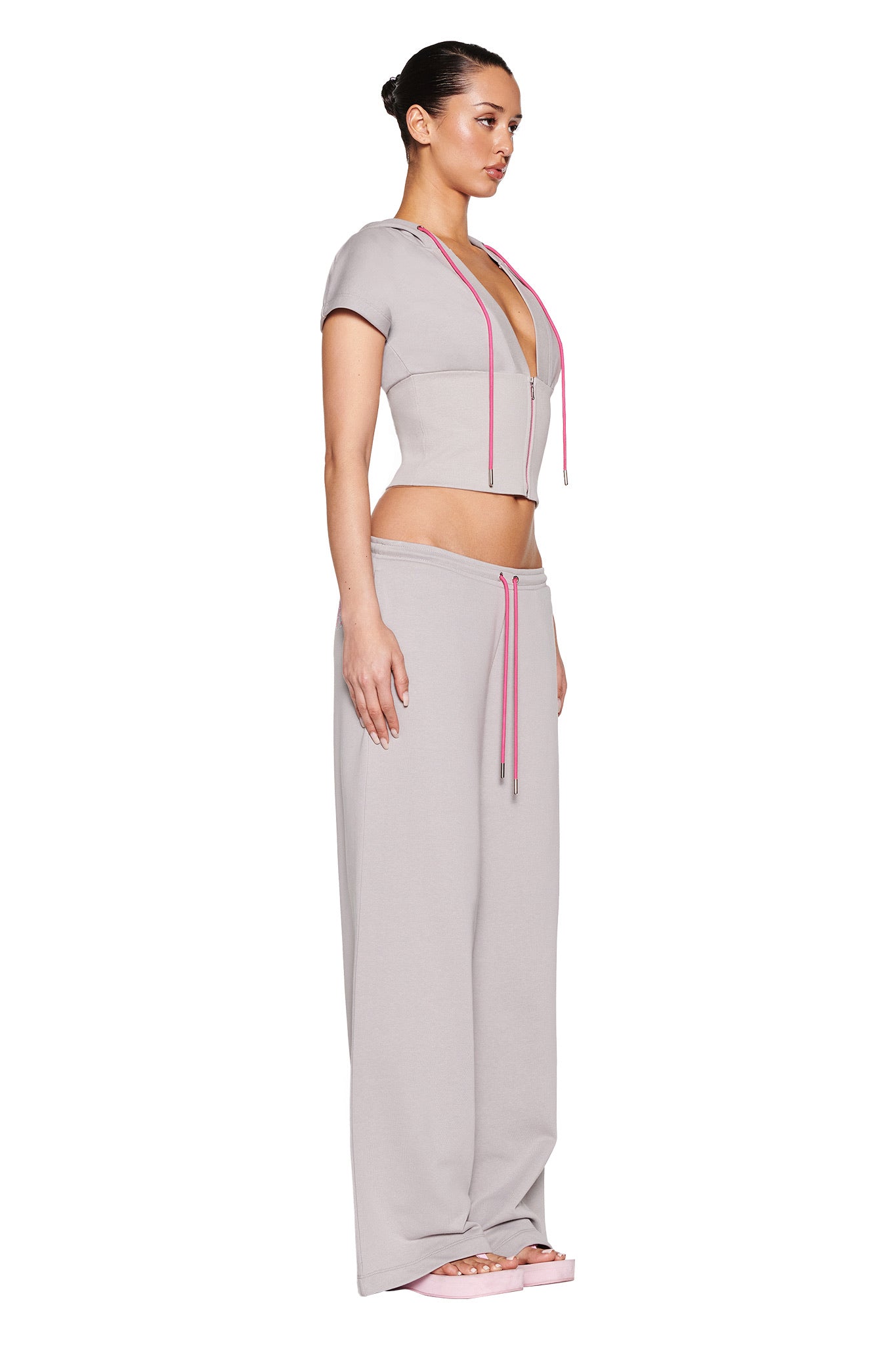 A person is in profile, donning the I.AM.GIA ARLO TRACK PANT - GREY : PINK two-piece with a cropped top featuring pink drawstrings and branding. The low-rise, wide-leg pants pair well with a neat bun and platform sandals, all highlighted against a plain white background.