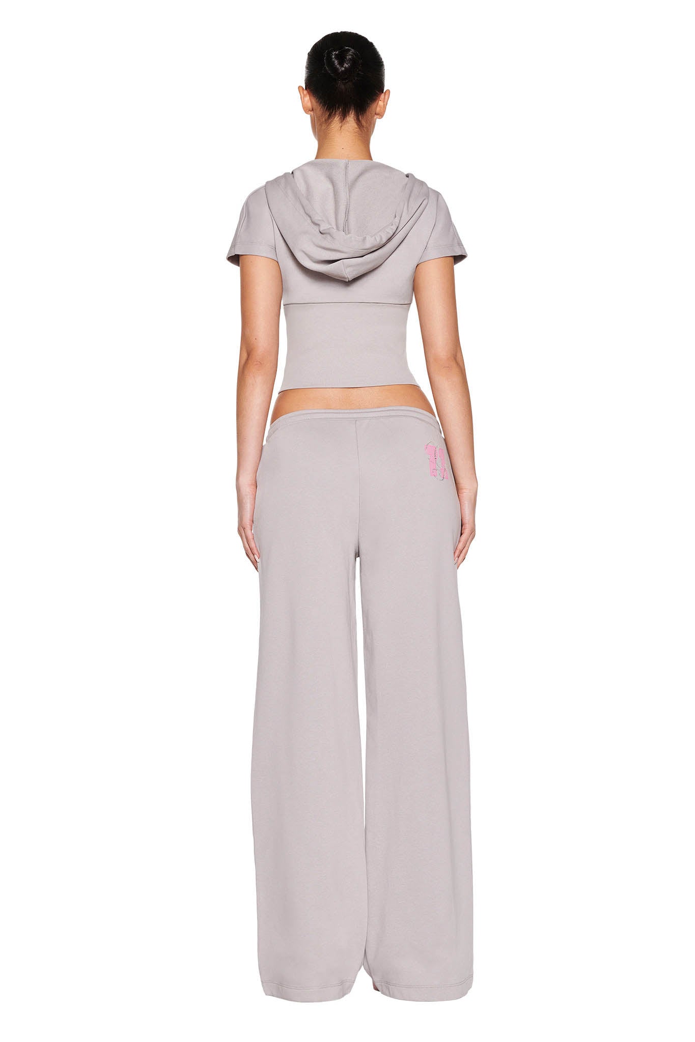 Rear view of a person wearing the ARLO TRACK PANT in grey and pink by I.AM.GIA, featuring a light gray, short-sleeved hooded crop top with an I.AM.GIA print. The outfit is casual, complementing the low-rise, wide-leg track pants as the person stands against a white background.
