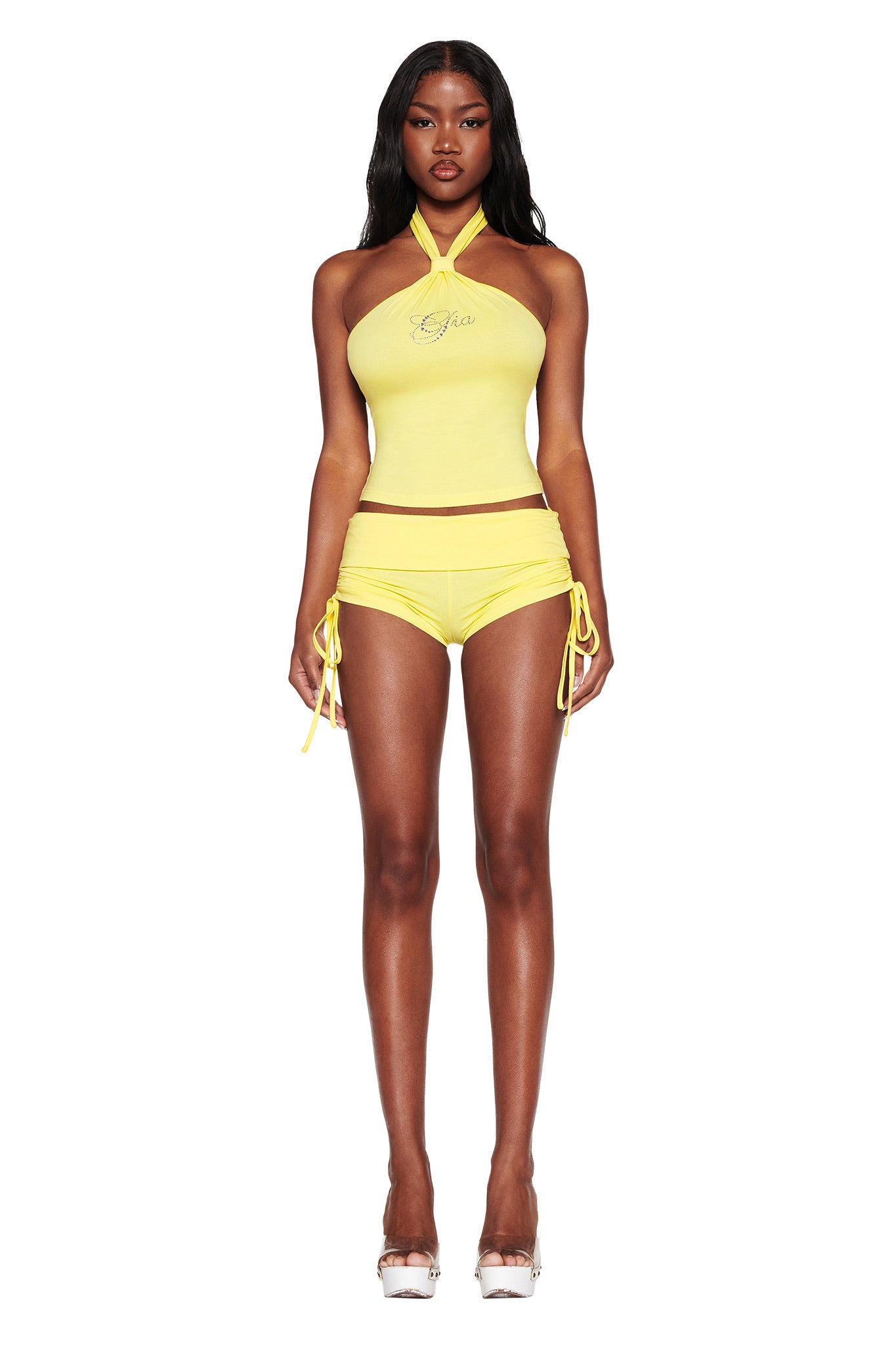 A person stands confidently in the I.AM.GIA ROWAN TOP - YELLOW with a sparkling diamanté logo and matching side-tie shorts. They have long hair, wear white high heels, all against a plain white background.