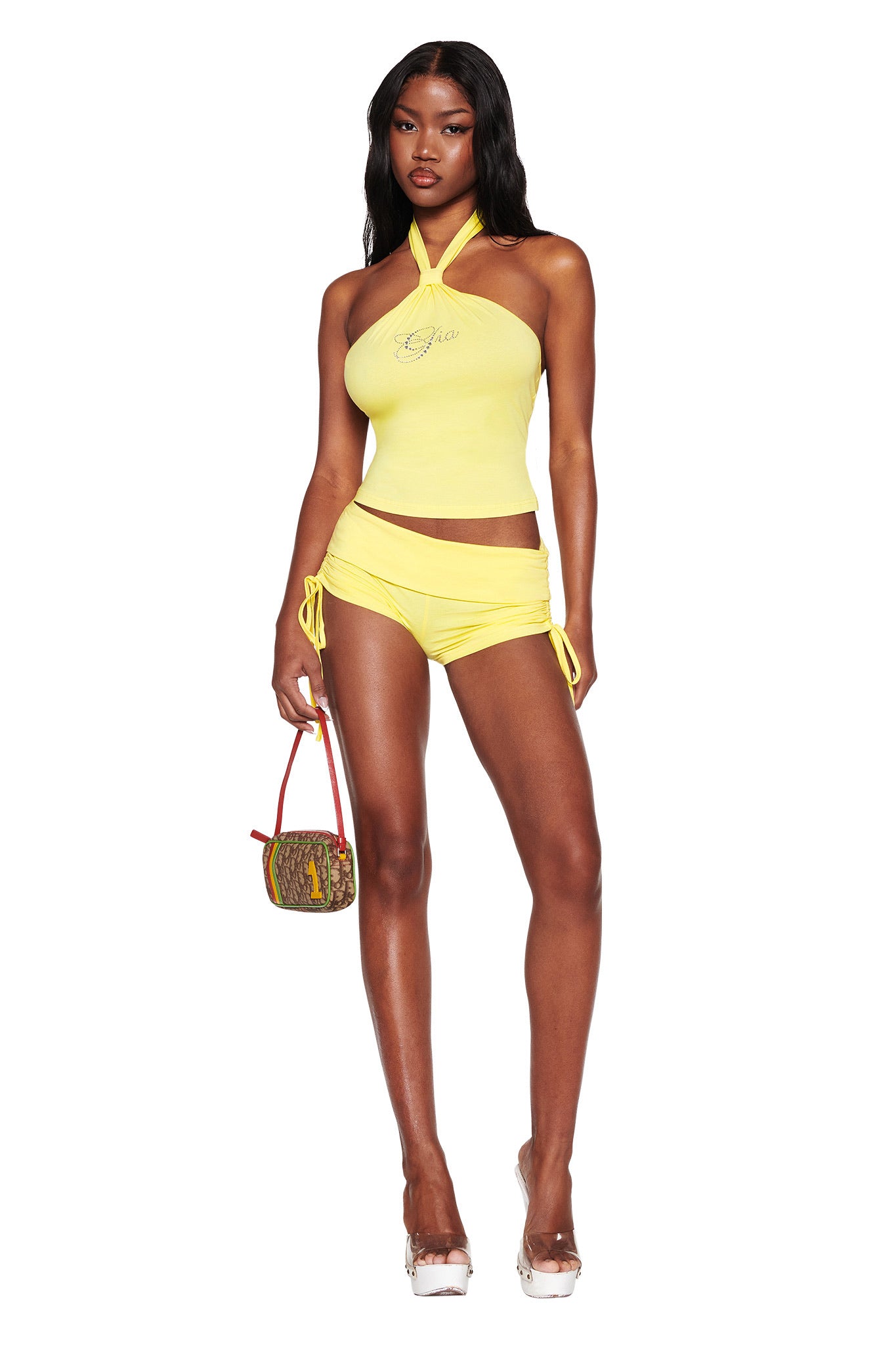A person stands against a white background wearing the I.AM.GIA ROWAN TOP in yellow and matching drawstring shorts, paired with a small brown handbag and clear high-heeled shoes for a chic look.