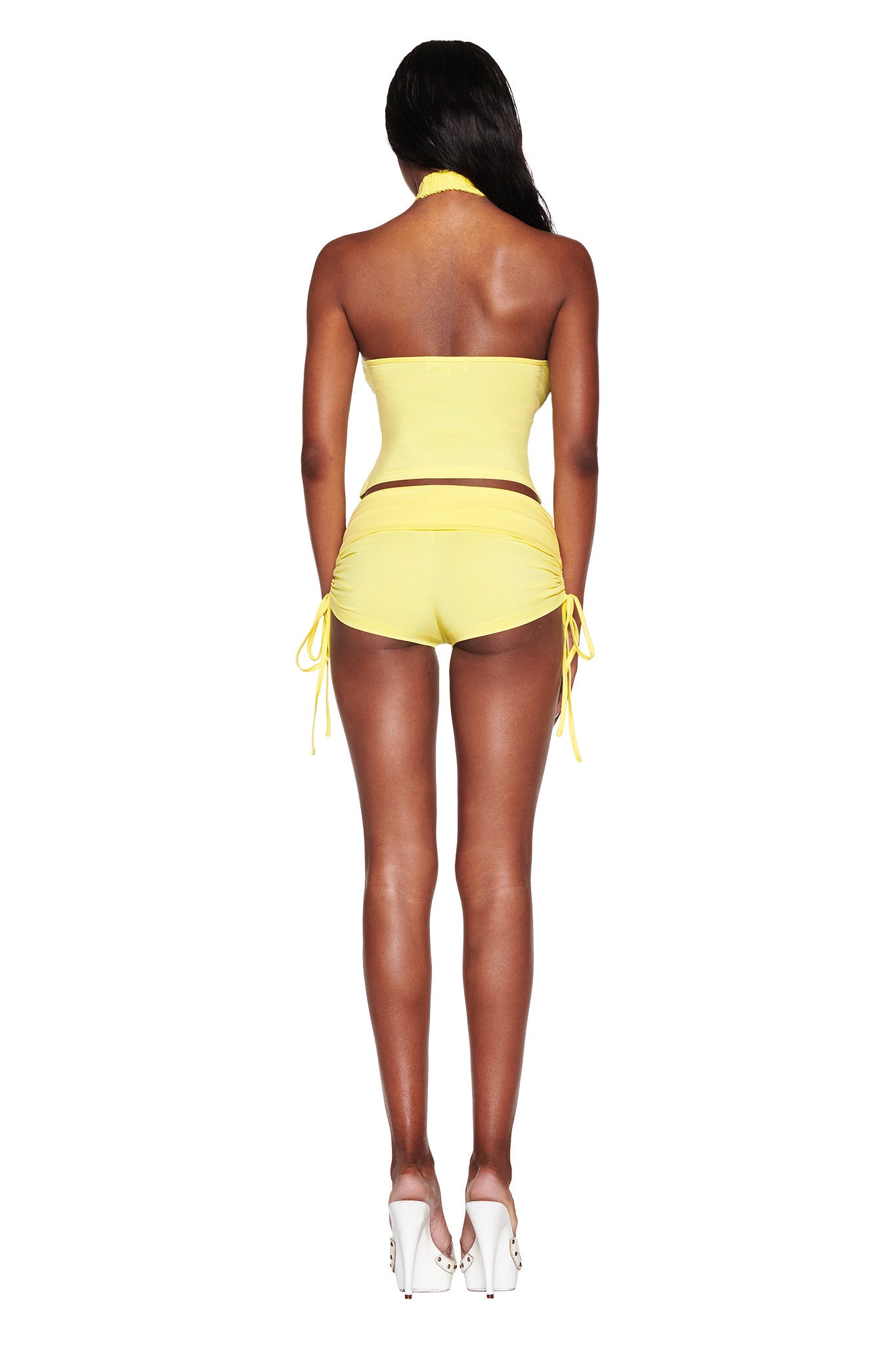 A person is turned away, sporting the I.AM.GIA ROWAN TOP in baby yellow with matching side-tie shorts. High heels and a subtle diamanté logo complete the chic look against a plain white background.