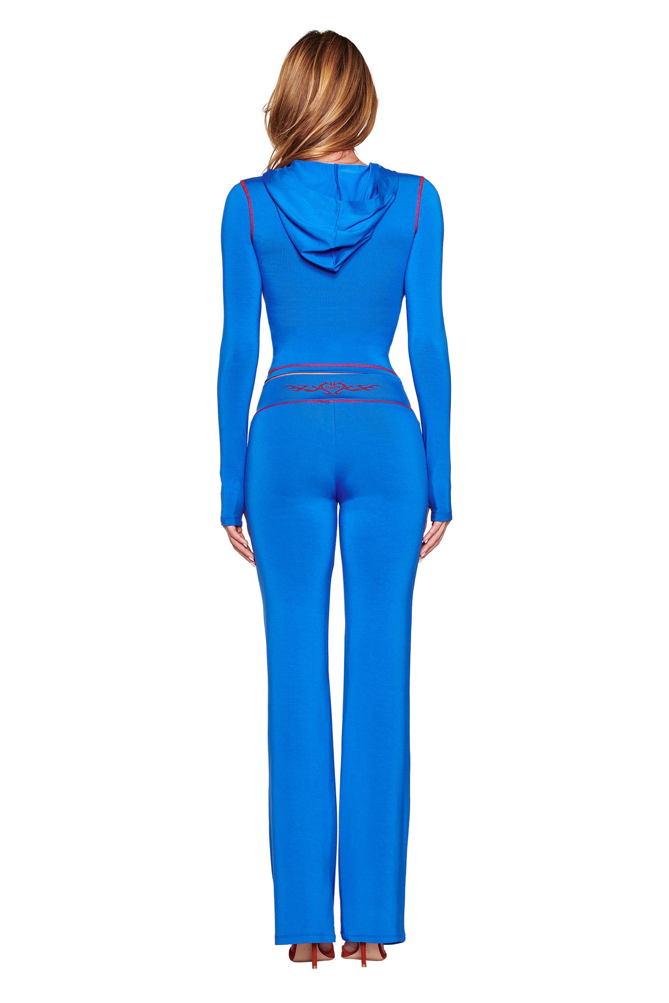 Rear view of a person in the I.AM.GIA BLARE TRACKPANT 2.0 - BLUE, showcasing vibrant royal blue with red accents and a signature design. The outfit features a hooded top and flared pants that perfectly complement their bodycon silhouette against a plain white background, with long light brown hair.