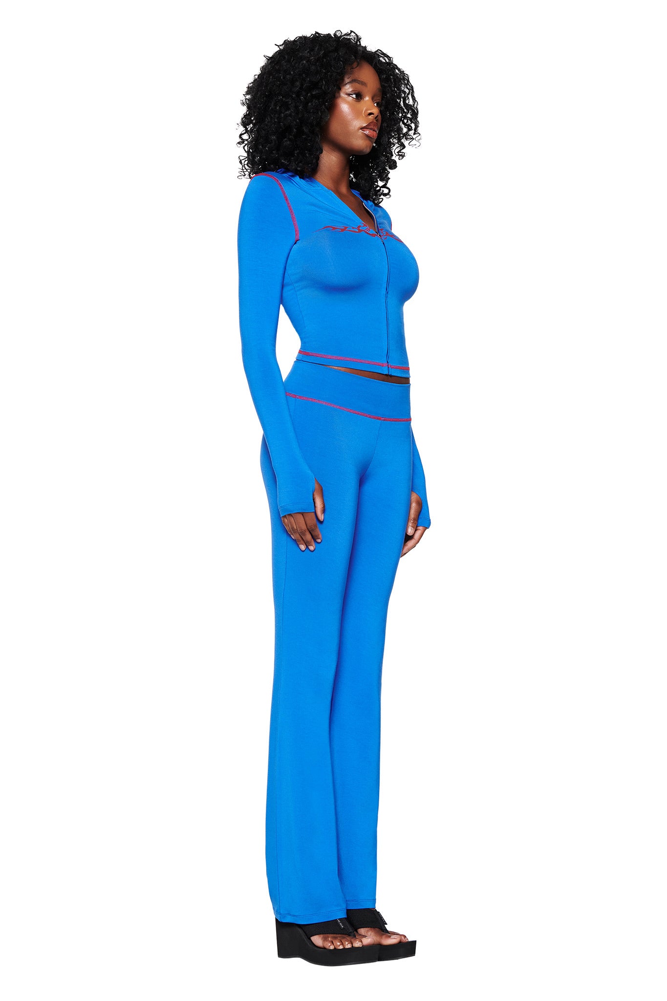 A woman with curly hair wears a fitted I.AM.GIA two-piece, the Blare Trackpant 2.0 in Royal Blue, featuring a bodycon design and red stitching. She poses against a white background in her long-sleeve zip-up top and black platform sandals, gazing to the side.