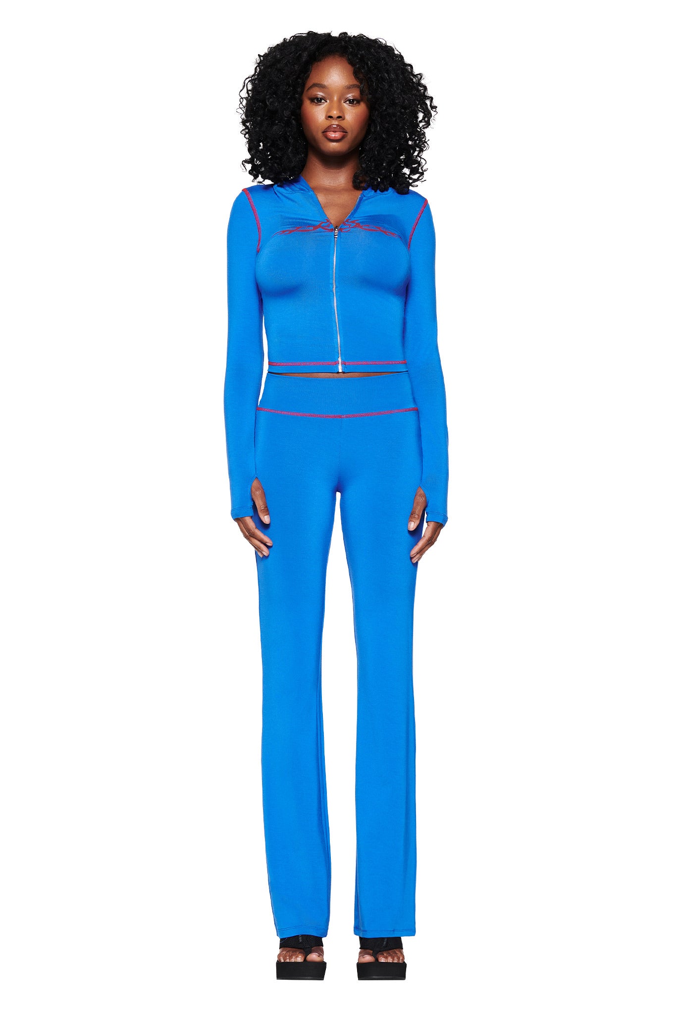 A woman in a royal blue zip-up top and I.AM.GIA's BLARE TRACKPANT 2.0 stands facing forward, showcasing her bodycon silhouette with red stitched edges. Her curly black hair complements the ensemble, paired with black platform shoes against a plain white backdrop.