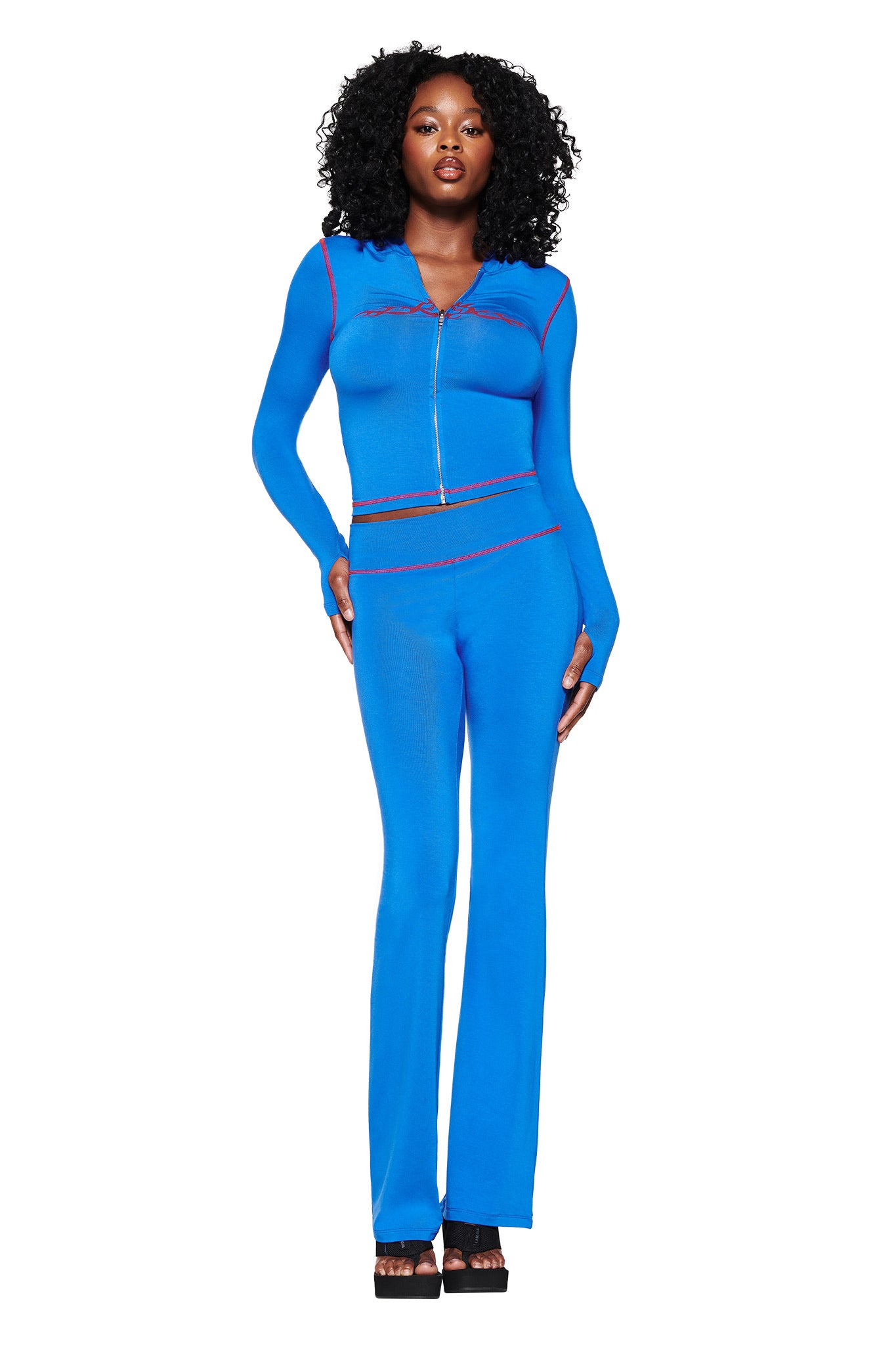 A person with curly hair wears a fitted royal blue outfit in a bodycon silhouette, featuring a long-sleeve zip-up top and flared pants like the I.AM.GIA BLARE TRACKPANT 2.0. They pair it with black platform shoes against a plain white background.
