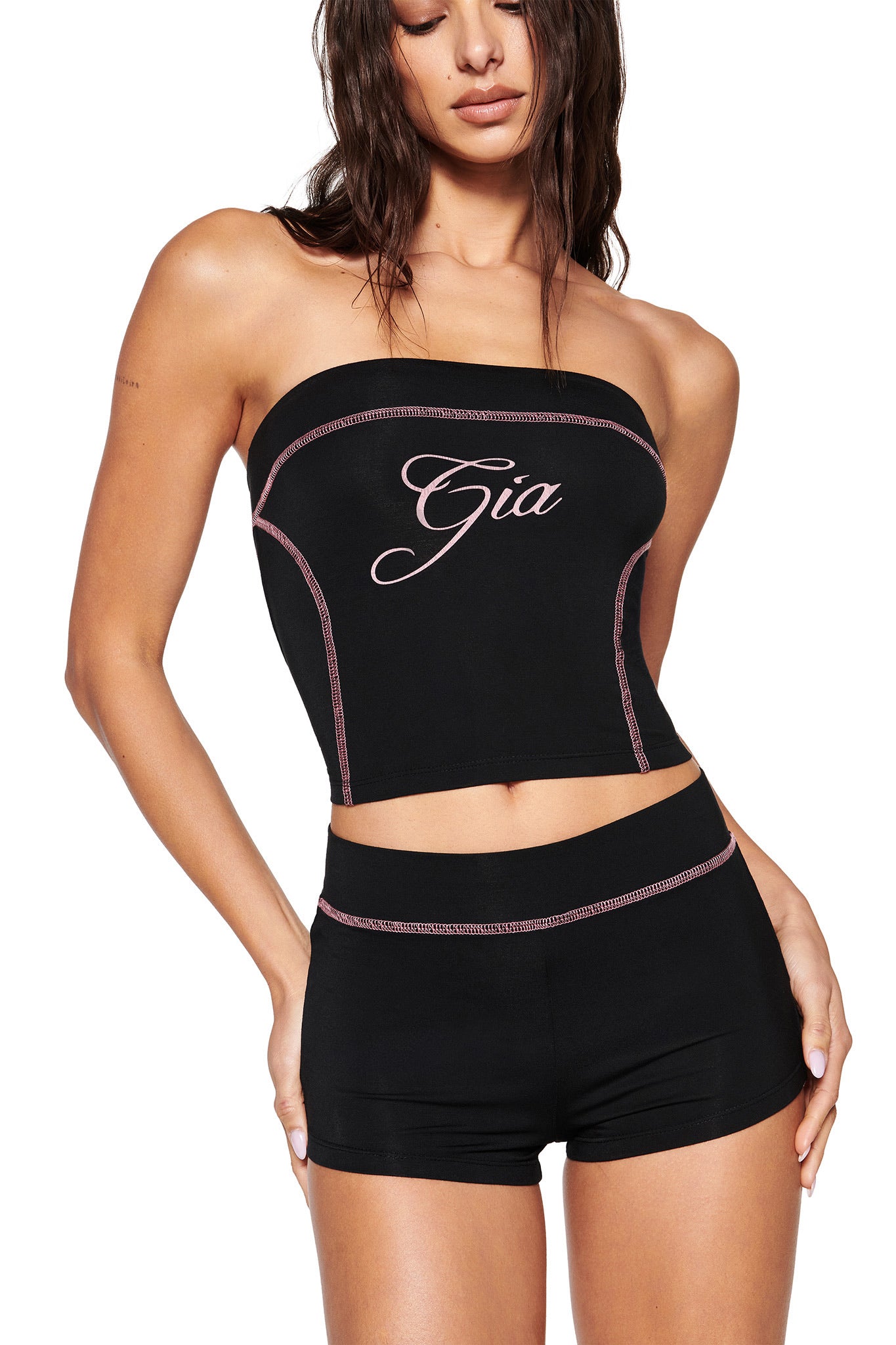 A person wears the BLARE SHORT - BLACK from I.AM.GIA, styled with a black strapless top featuring pink "Gia" script and matching pink-stitched bodycon shorts. Their long, dark, wet-look hair enhances this bold and stylish look.