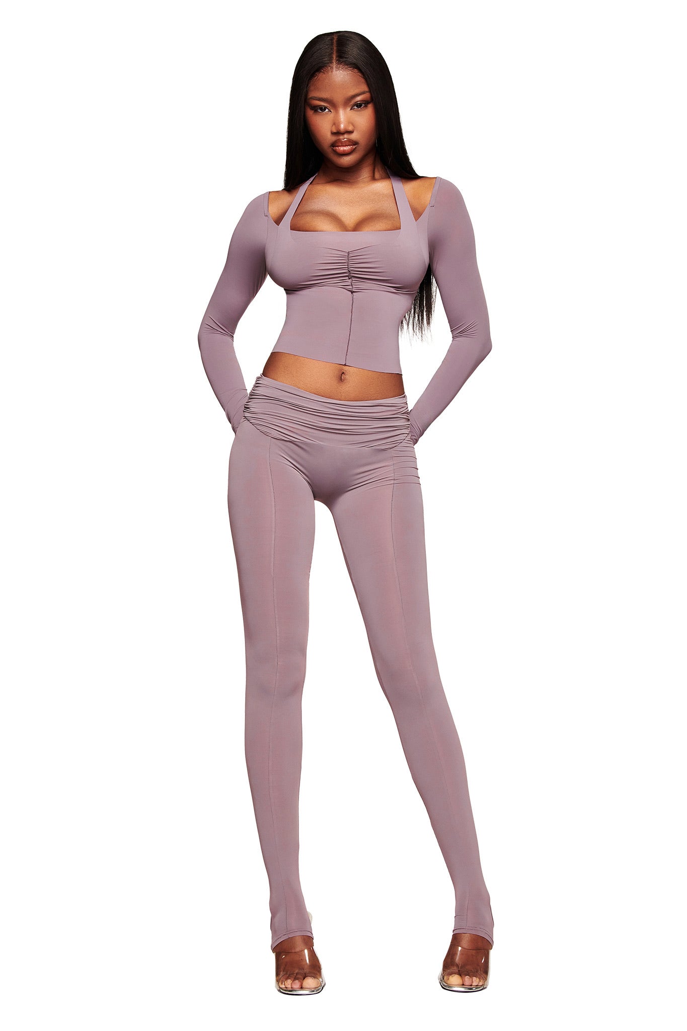 A woman stands confidently in the I.AM.GIA NERINA LEGGING in dusty lilac, featuring a fitted, high-waisted full-length legging paired with a cropped top and clear heels against a white background.