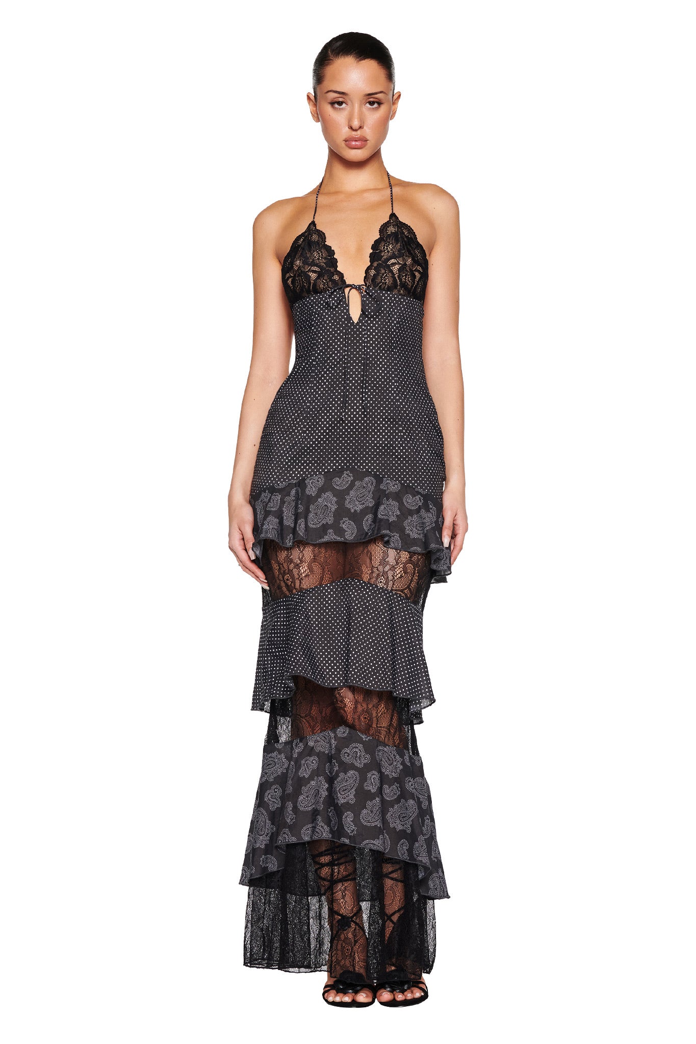 A woman poses against a white background wearing I.AM.GIA's PEARL MAXI DRESS - BLACK : POLKA DOT, showcasing its long, layered design with non-stretch lace and polka dot patterns. The elegant dress features a halter neck and tiered details, paired with black heels.