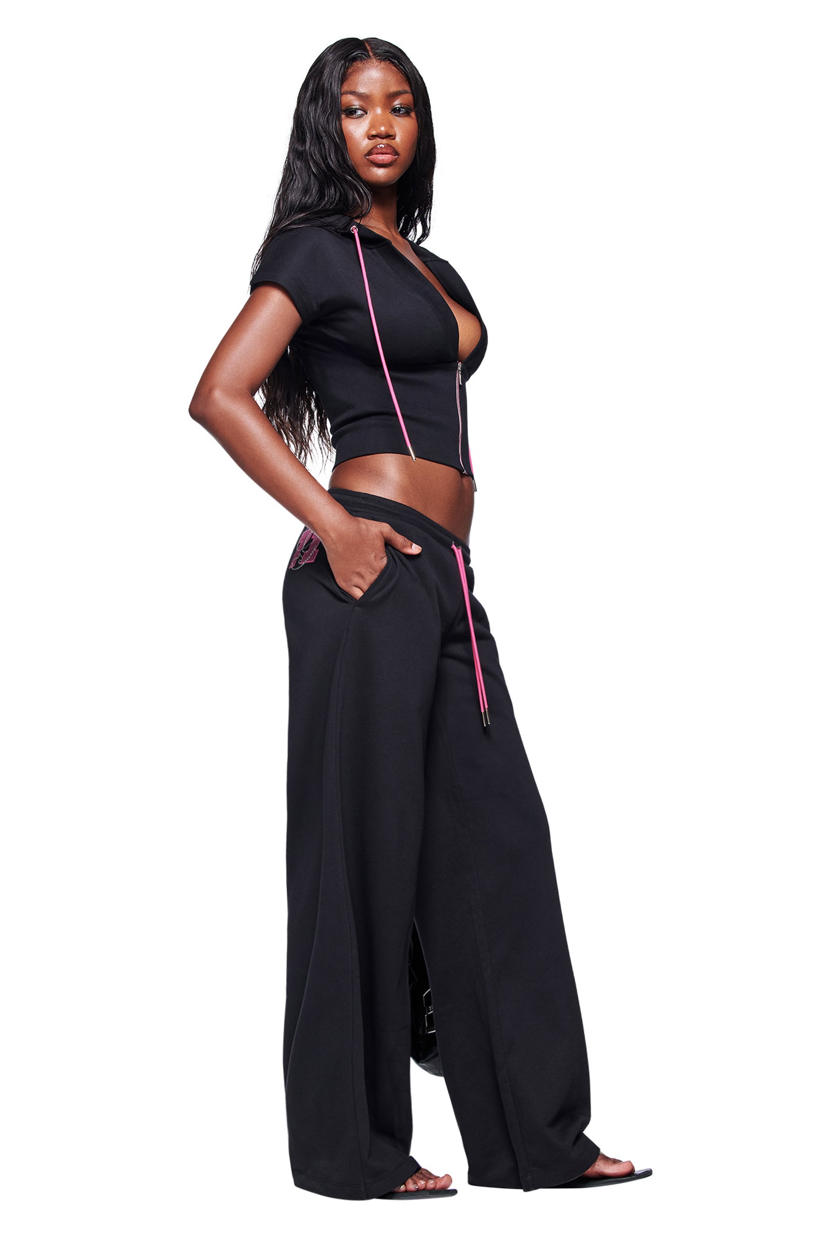 A woman with long wavy hair poses confidently against a white background, wearing a black short-sleeve crop top with pink strings and the I.AM.GIA ARLO TRACK PANT in black, with one hand in her pocket.