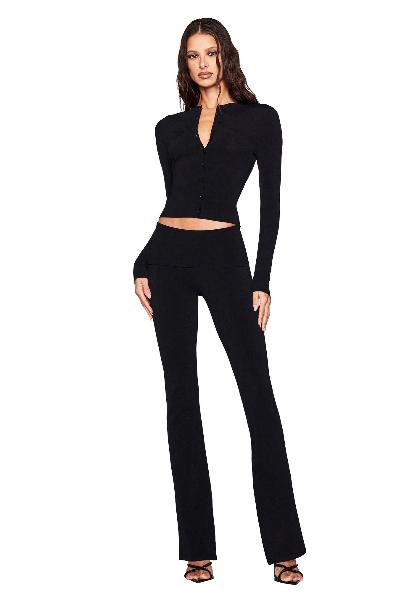 A woman wearing I.AM.GIA's AUDRINA PANT 2.0 in black, featuring a chic foldover waistband, pairs it with a black long-sleeve buttoned top and strappy heels. Her right hand on her hip and wavy hair enhance her confident bodycon fit ensemble on a white background.