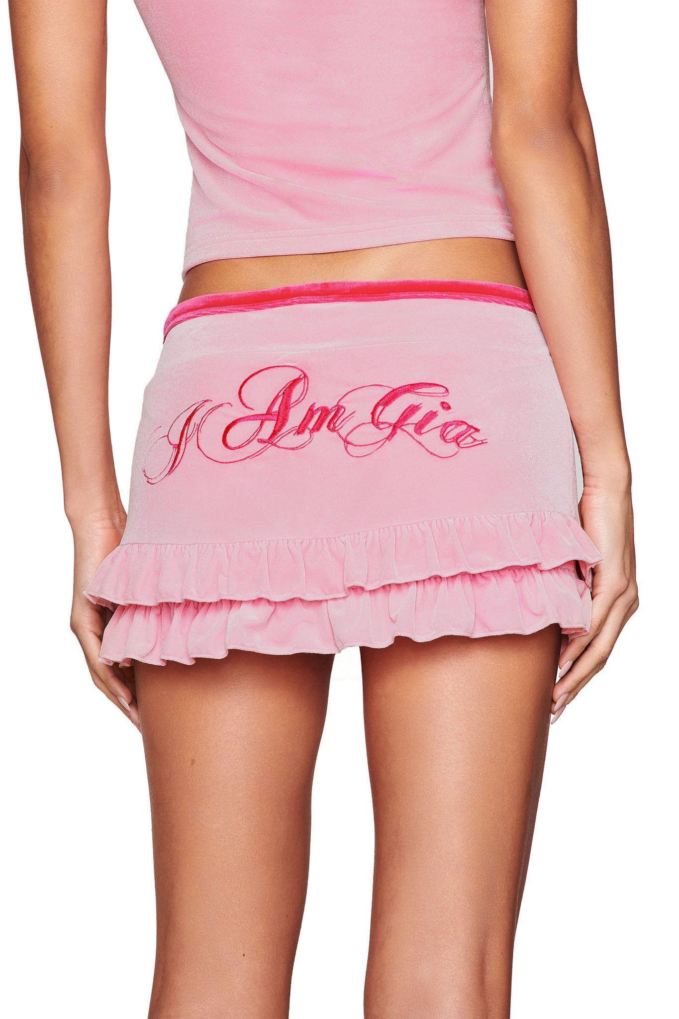 A person is seen from behind wearing a pink Michelle Velour Mini Skort, featuring red embroidered script and a low-rise waist, paired with a matching top. The skort showcases the brand name "I.AM.GIA.