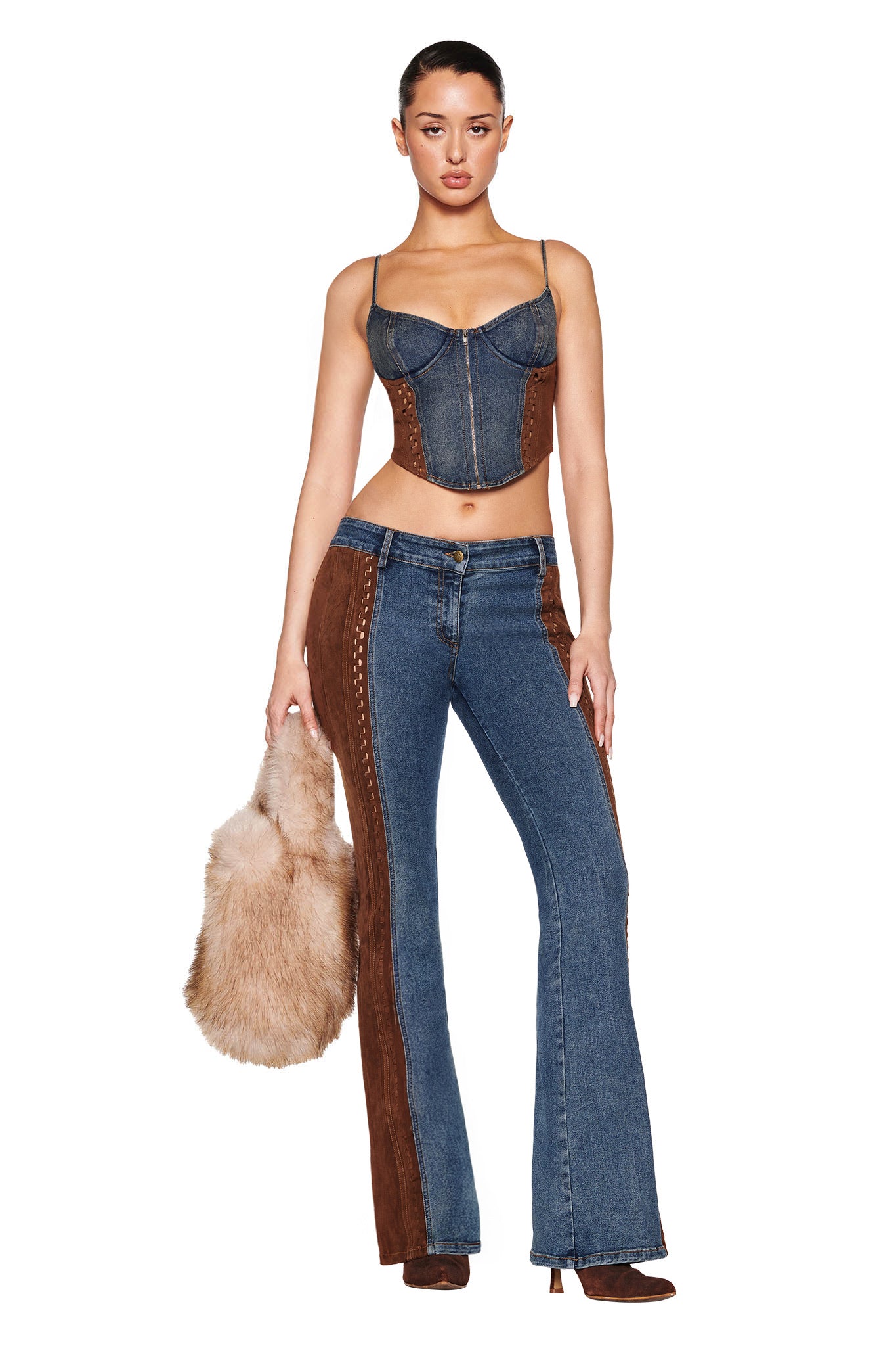 A woman in I.AM.GIA's FORREST PANT - BLUE: MEDIUM WASH and a denim corset top holds a furry beige handbag. Her hair is tied back, and she wears brown boots. The outfit, featuring mid-rise flare style with brown suede contrast panels, exudes a vintage washed retro vibe.