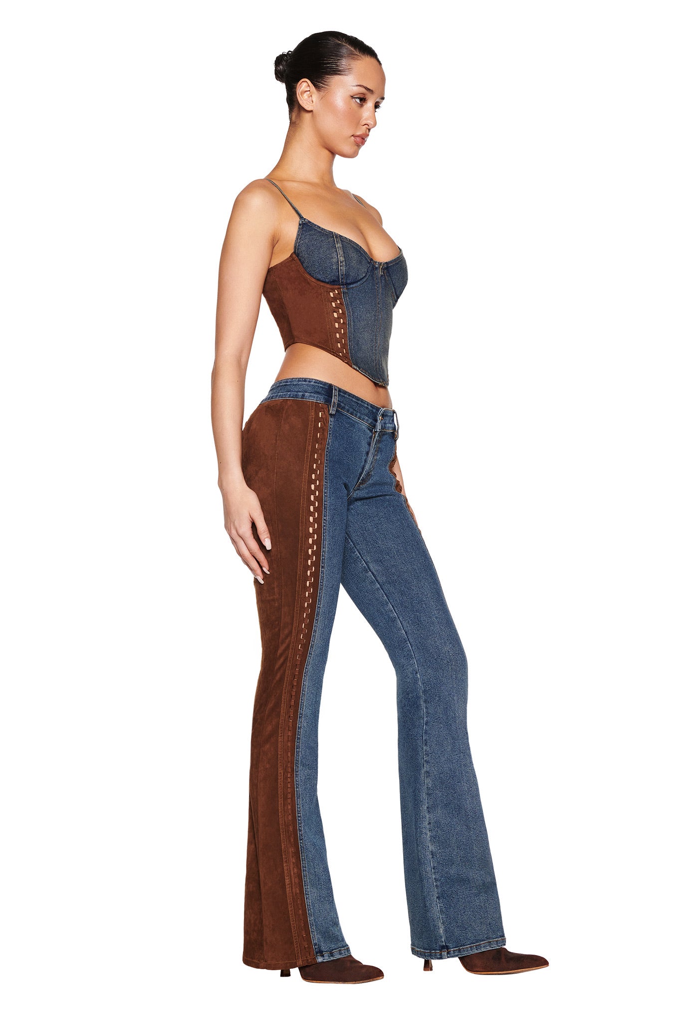 A person poses against a plain white background, wearing I.AM.GIA's FORREST BUSTIER in medium wash blue denim with a brown corset design, paired with matching flared pants featuring unique stitching, suede contrast panels, and adjustable straps.
