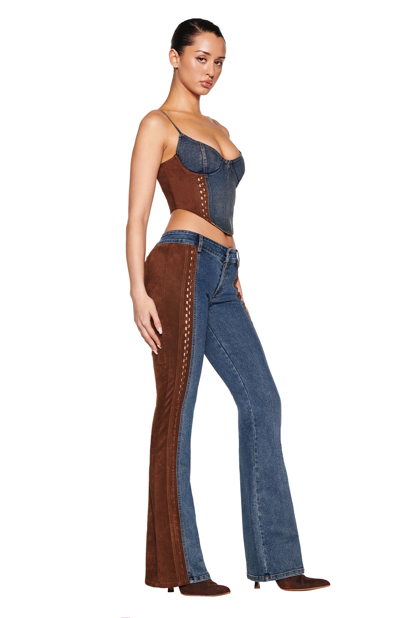 A person confidently poses in I.AM.GIA's FORREST PANT in medium wash, a vintage-inspired denim featuring suede contrast panels. The outfit includes a corset-style top and flared jeans, completed with brown heels against a white background.