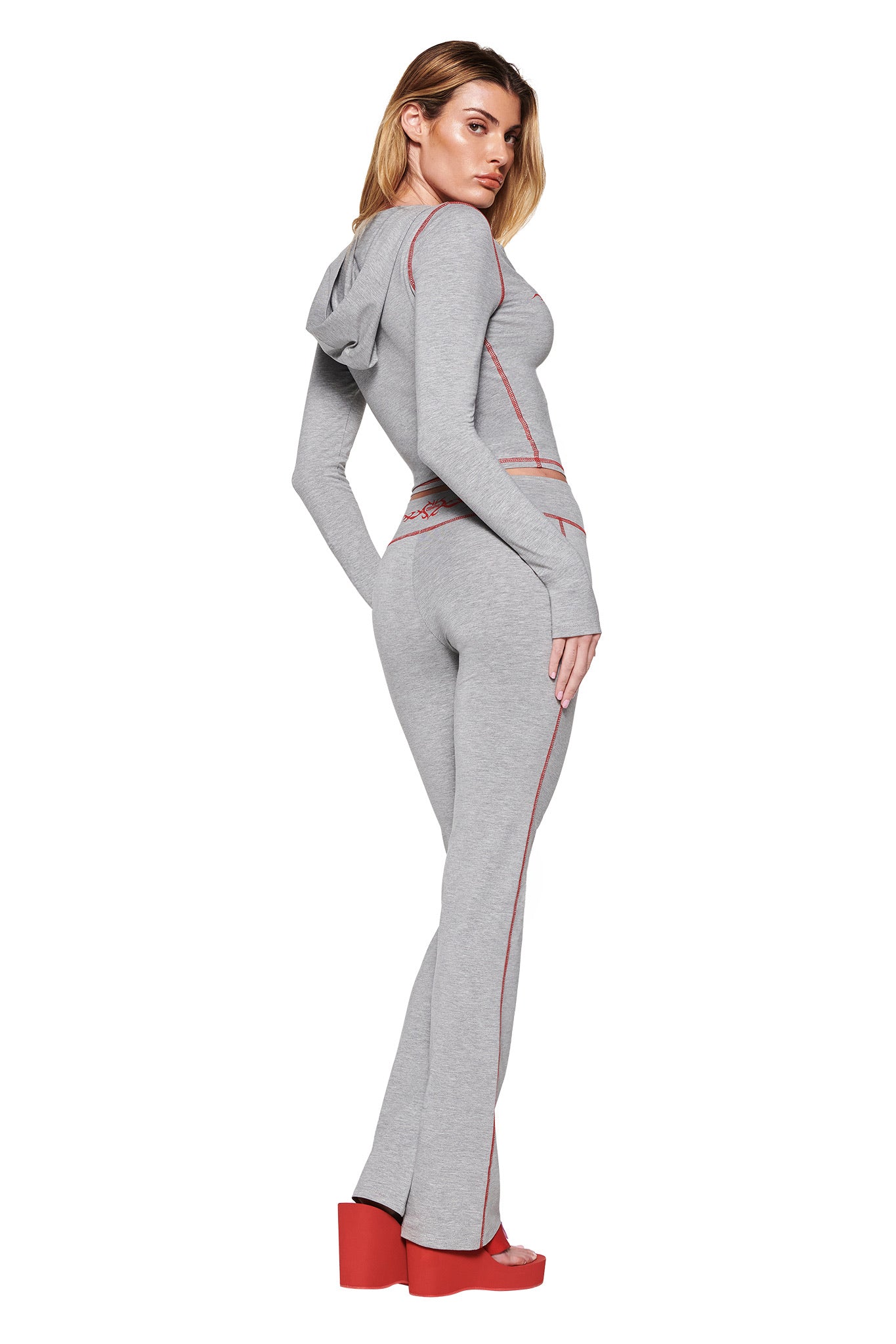 A person modeling a snug Dove Grey Blare Hoodie from I.AM.GIA, featuring the GIA Flame logo and red accents, stands sideways. They wear flared pants and striking red platform shoes.