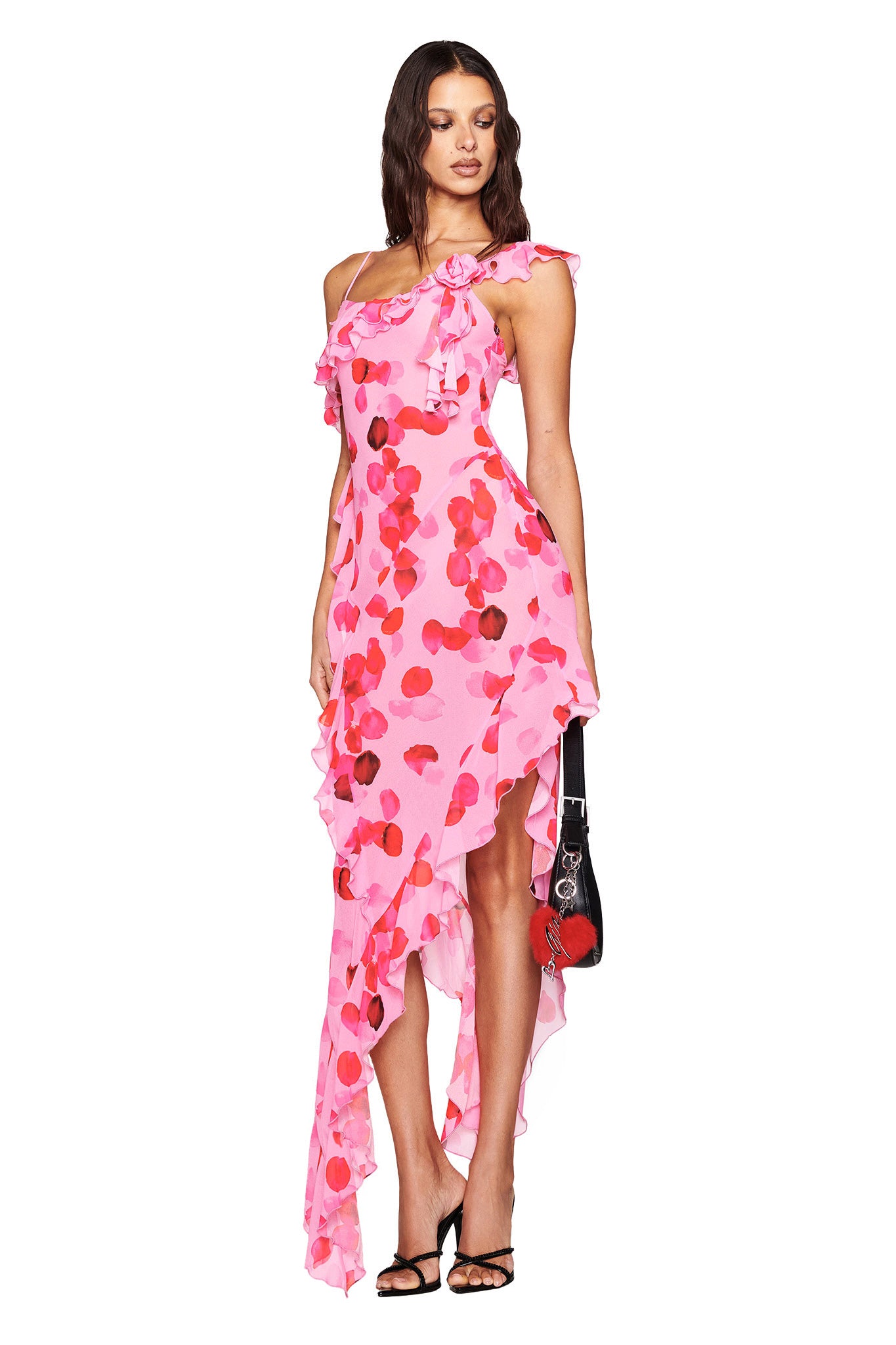 A woman in an I.AM.GIA JULIETTA MAXI DRESS - PINK: PEACH PRETTY PETAL with ruffles and a high-low hemline. She holds a black handbag with red detail and wears black strappy heels. Her hair is wavy, and the dress features an asymmetrical design with a single shoulder strap.