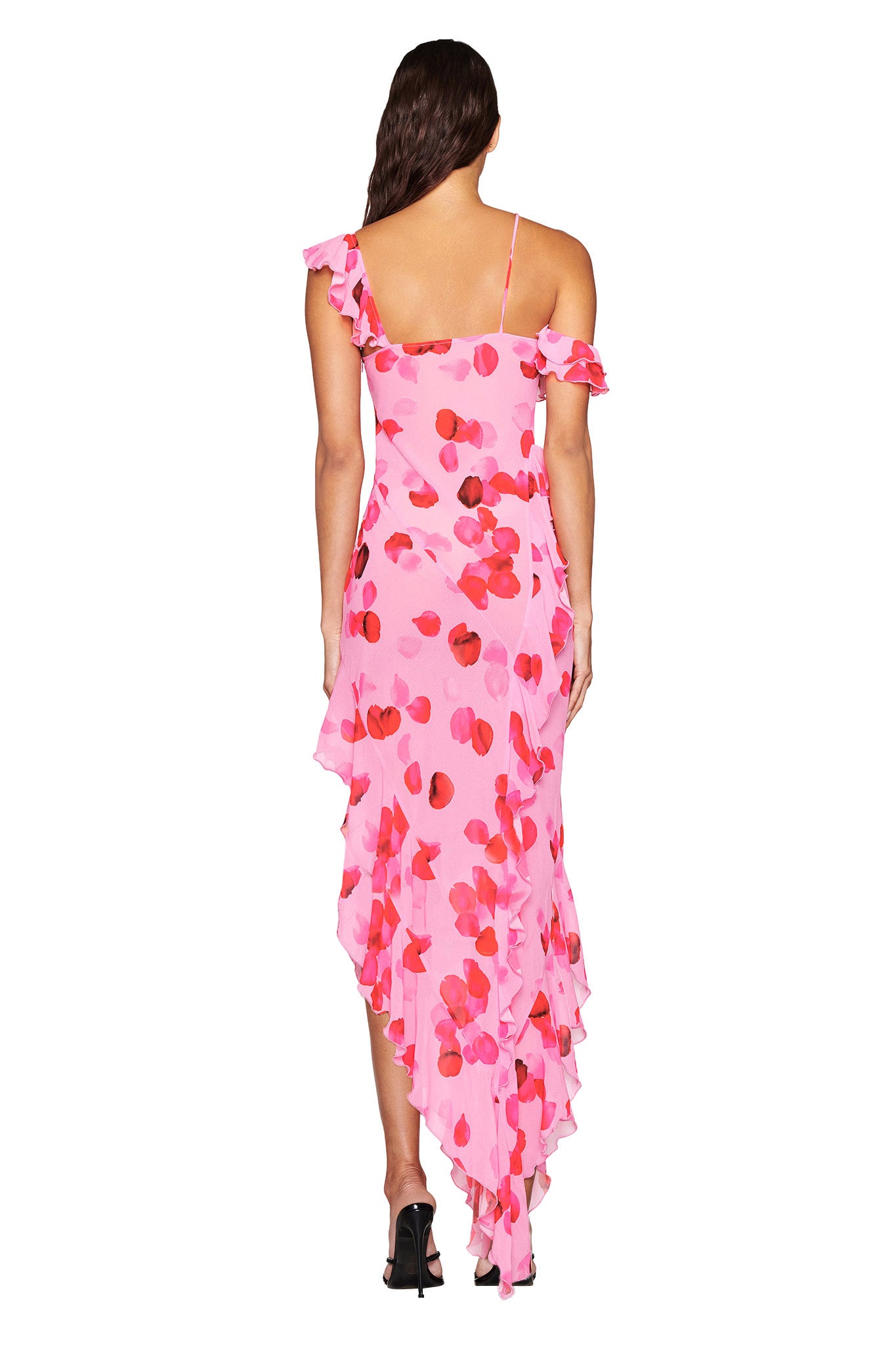 A woman in the JULIETTA MAXI DRESS from I.AM.GIA, featuring a pink design with pretty peach petal print, is facing away. The dress highlights ruffled off-the-shoulder sleeves and an asymmetrical hem, paired perfectly with black high-heeled shoes.
