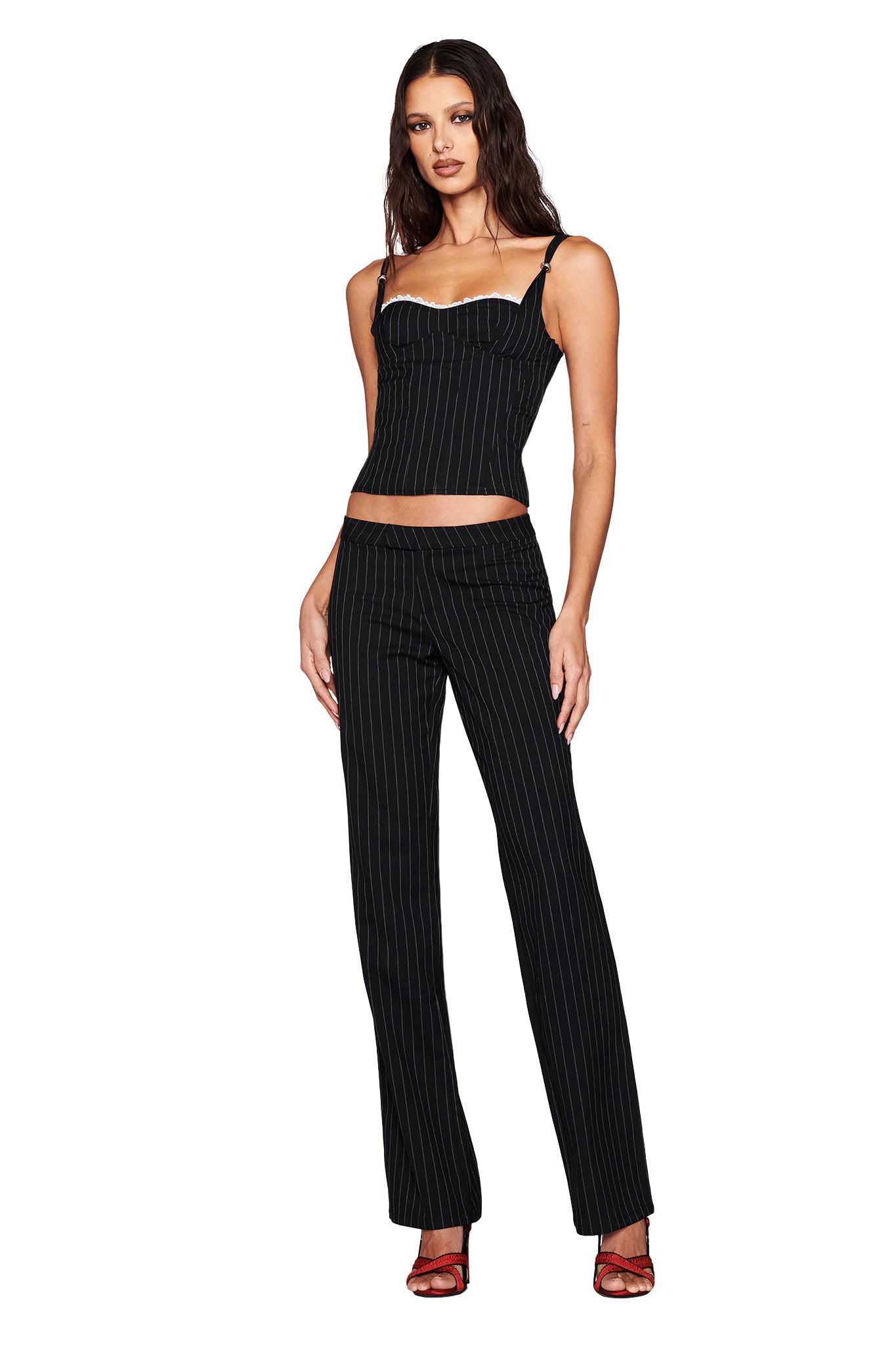 A person is wearing the I.AM.GIA MARRION CORSET in black, paired with matching pinstripe pants, featuring lace trim. They have long, wavy hair and are standing relaxed. Their shoes are red high heels on a white background.