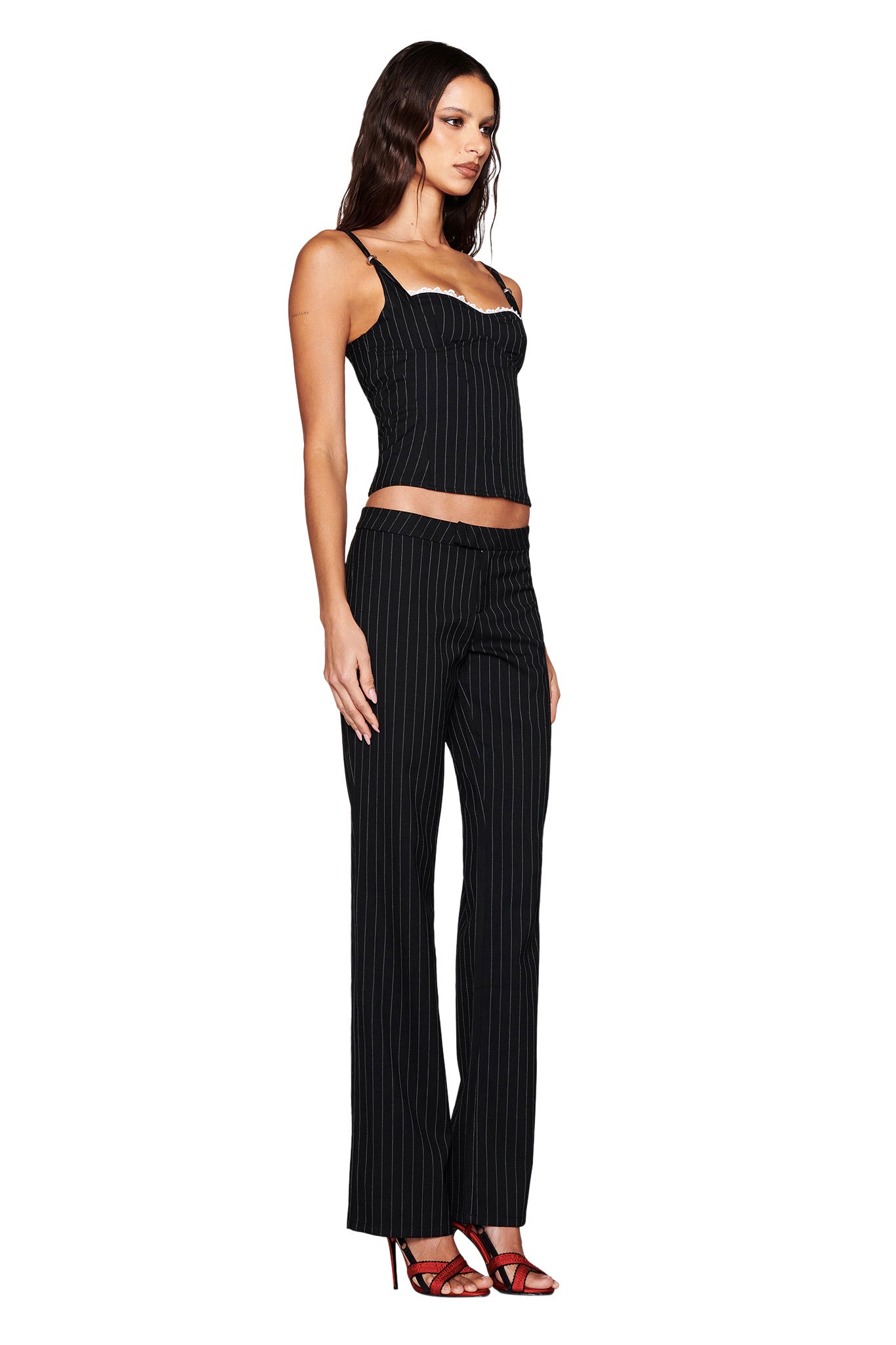 A woman in profile against a white background wears the I.AM.GIA MARRION CORSET - BLACK, featuring delicate lace, paired with matching flared pants and red strappy high heels.
