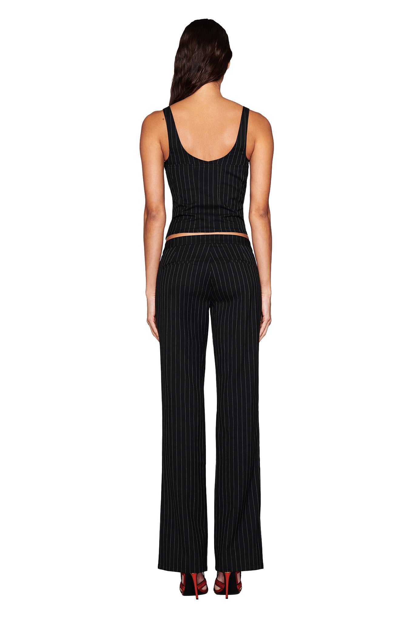 Rear view of a person with long dark hair wearing I.AM.GIA's MARRION CORSET in black, styled as a sleeveless pinstripe top paired with flared pants, standing elegantly against a white background.
