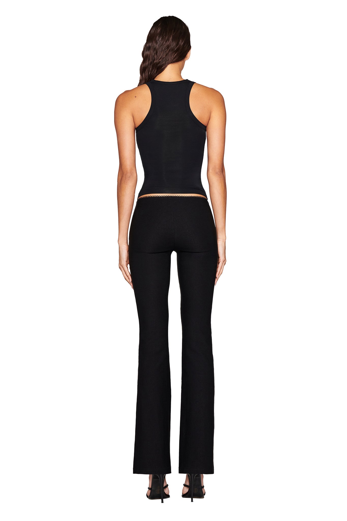 A person with long hair wears the I.AM.GIA AQUARIUS ZODIAC TANK - BLACK, black pants, and black heels; the sleek outfit stands out against the white background.