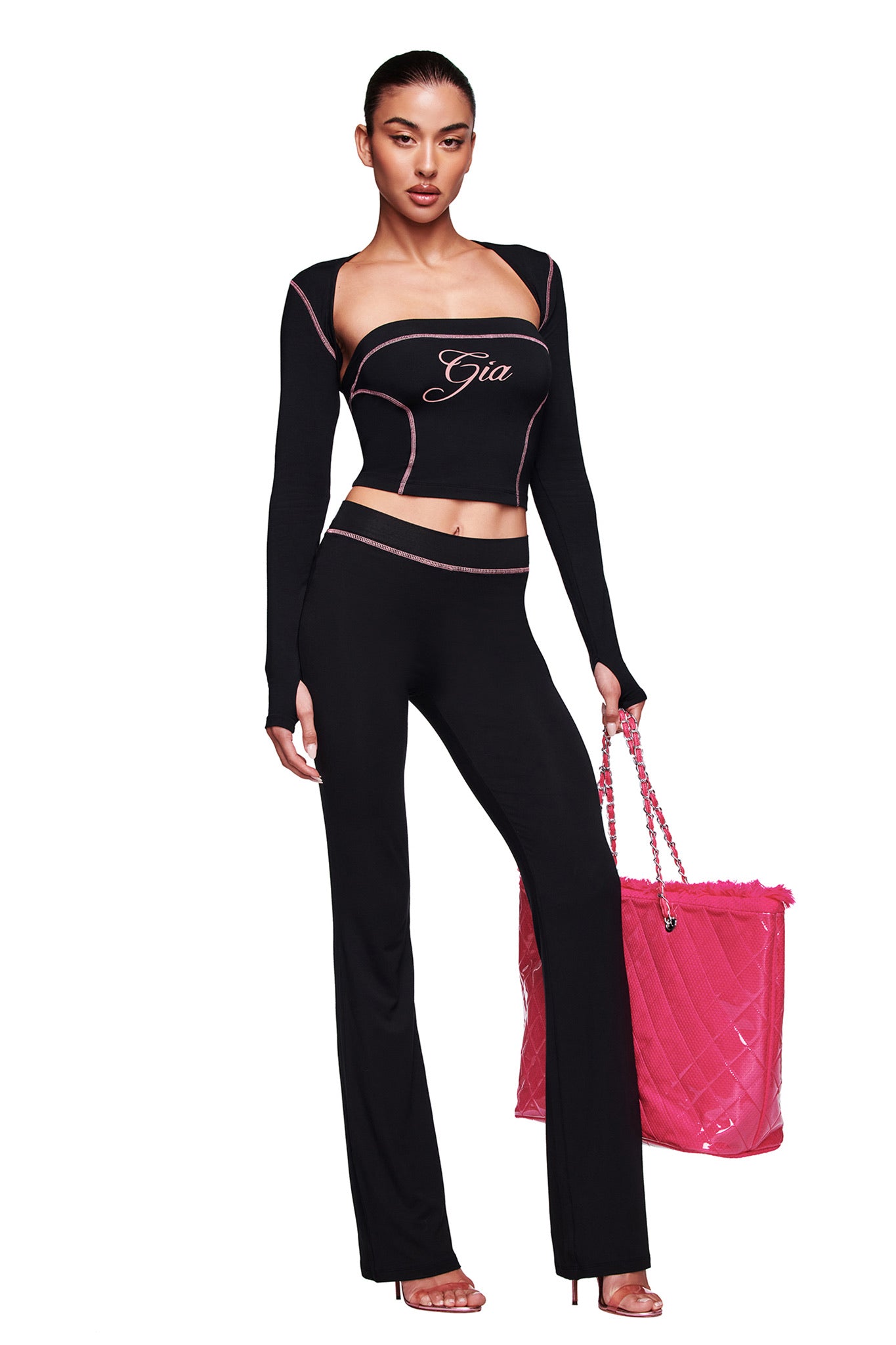 A woman models the BLARE BOOB TUBE in black by I.AM.GIA, a two-piece with pink trim and sporty princess seams. “Gia” appears on the top. She holds a large pink quilted chain bag and wears high heels against a plain white background.
